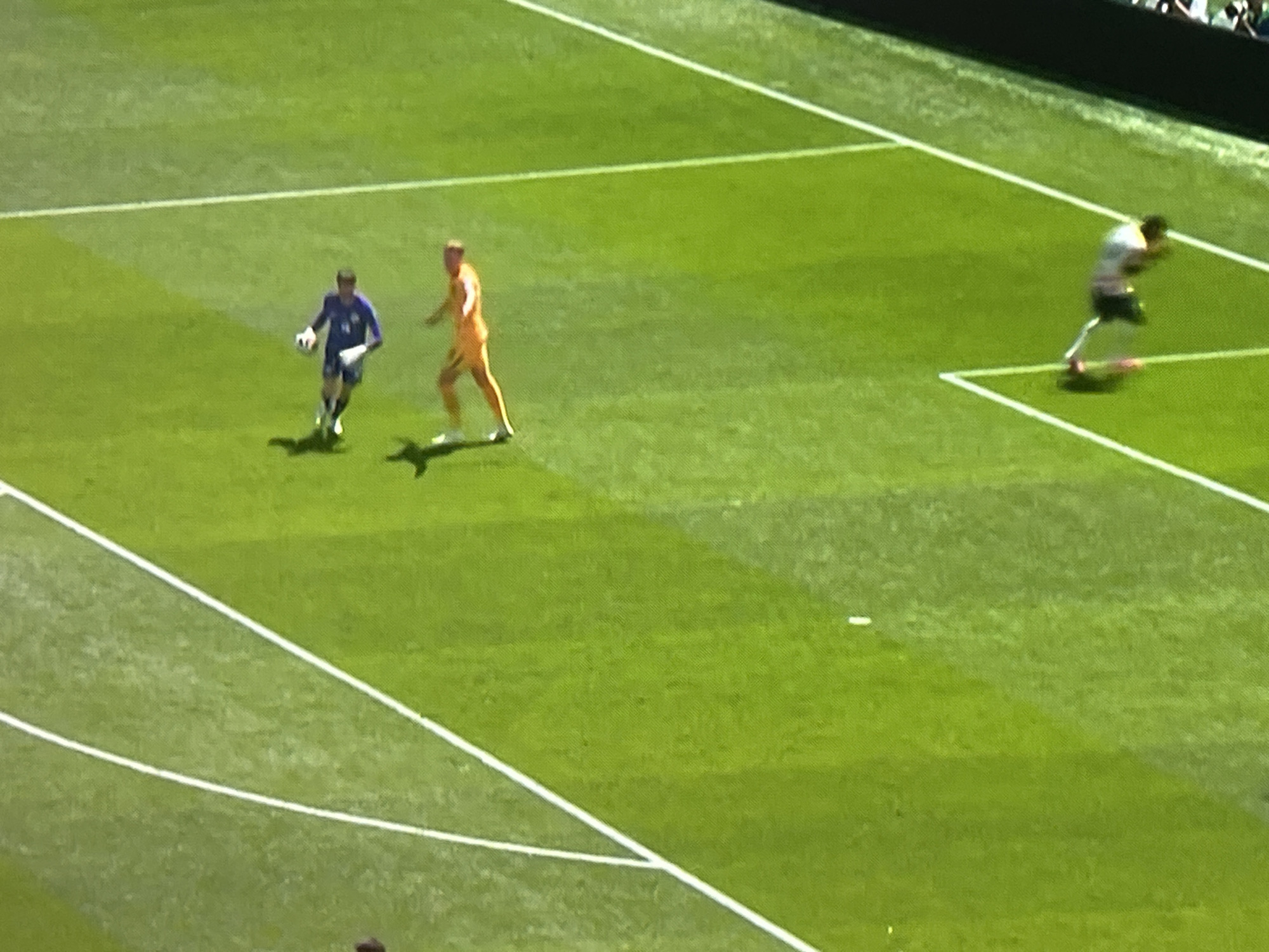 The image shows a wide shot of a soccer field with three individuals visible. In the foreground left, a person wearing a blue uniform is running with a soccer ball. To their right, another person in an orange uniform is also on the field. Further in the background right, a third individual in a white and black uniform is running. The field is green with white lines demarcating boundaries and penalty areas. The scene appears to be a sunny day, with bright lighting and visible shadows cast by the players. The overall setting is a well-maintained outdoor sports arena. No text is visible in the image. The activity taking place is a soccer game or practice. The focus is on the players and their movements on the field.