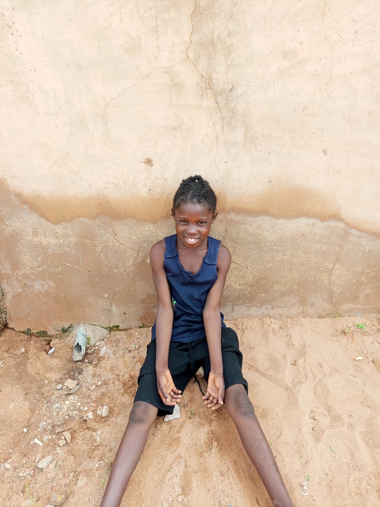 A young girl with dark skin and braided hair sits on sandy ground against a textured, beige wall in Tempure, Nigeria. She is wearing a navy blue sleeveless shirt and black shorts. Her legs are spread apart, and her arms rest on her knees, with her hands clasped. She smiles directly at the camera. The wall behind her shows signs of age and weathering, with cracks and a horizontal band of darker discoloration. The ground is composed of sand and small pebbles, with a piece of broken white plastic pipe visible near the left side of the frame.