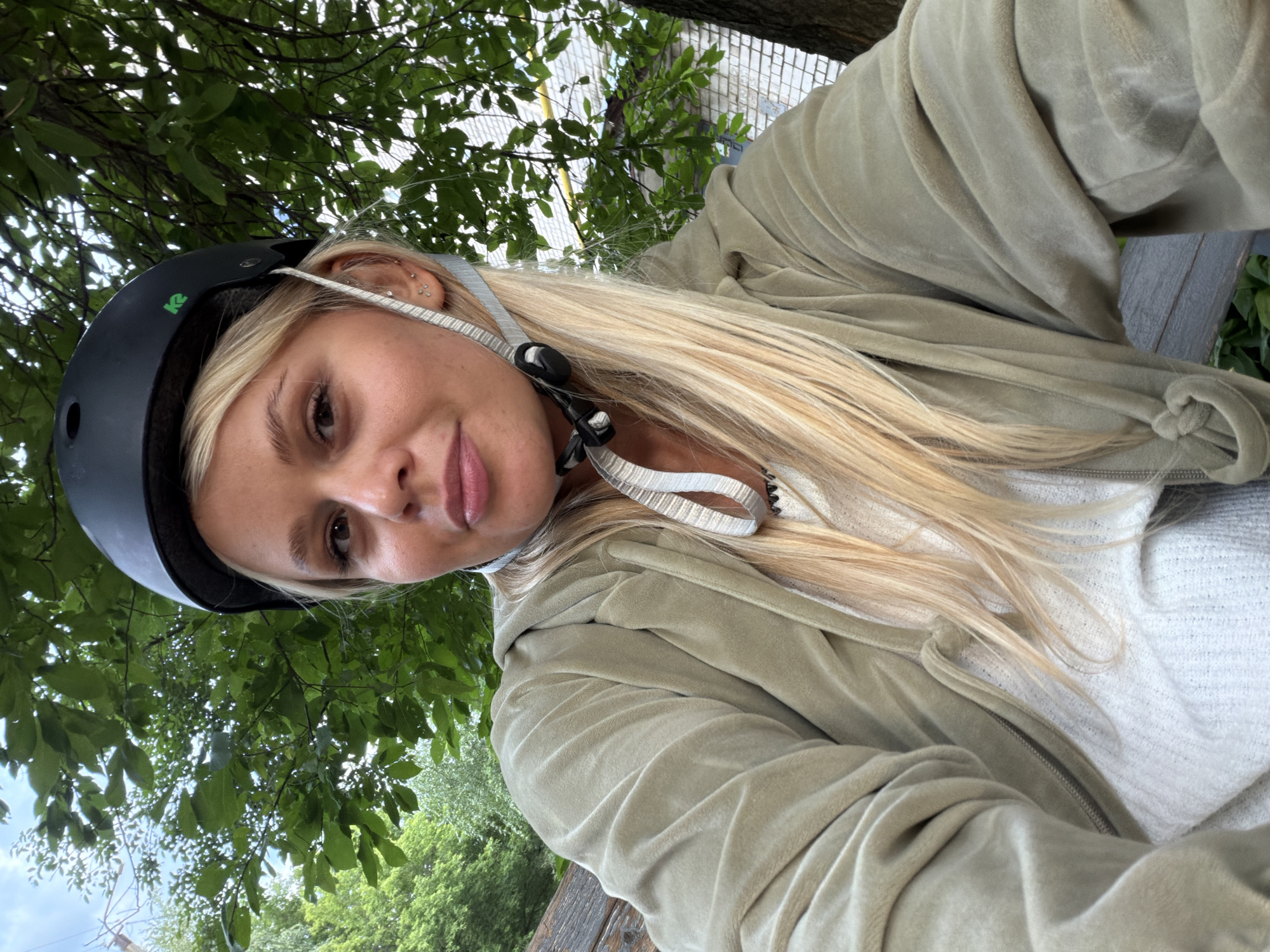 A young Caucasian woman with blonde hair and a light complexion is wearing a black helmet and a light green velour zip-up hoodie over a white textured sweater. She is outdoors, seated on a wooden bench under a tree with lush green leaves. In the background, a brick building is partially visible. The woman is looking at the camera and smiling slightly. Her hair is long and falls over her shoulders. The scene appears to be in a park or outdoor recreational area in Sarapul, Russia.