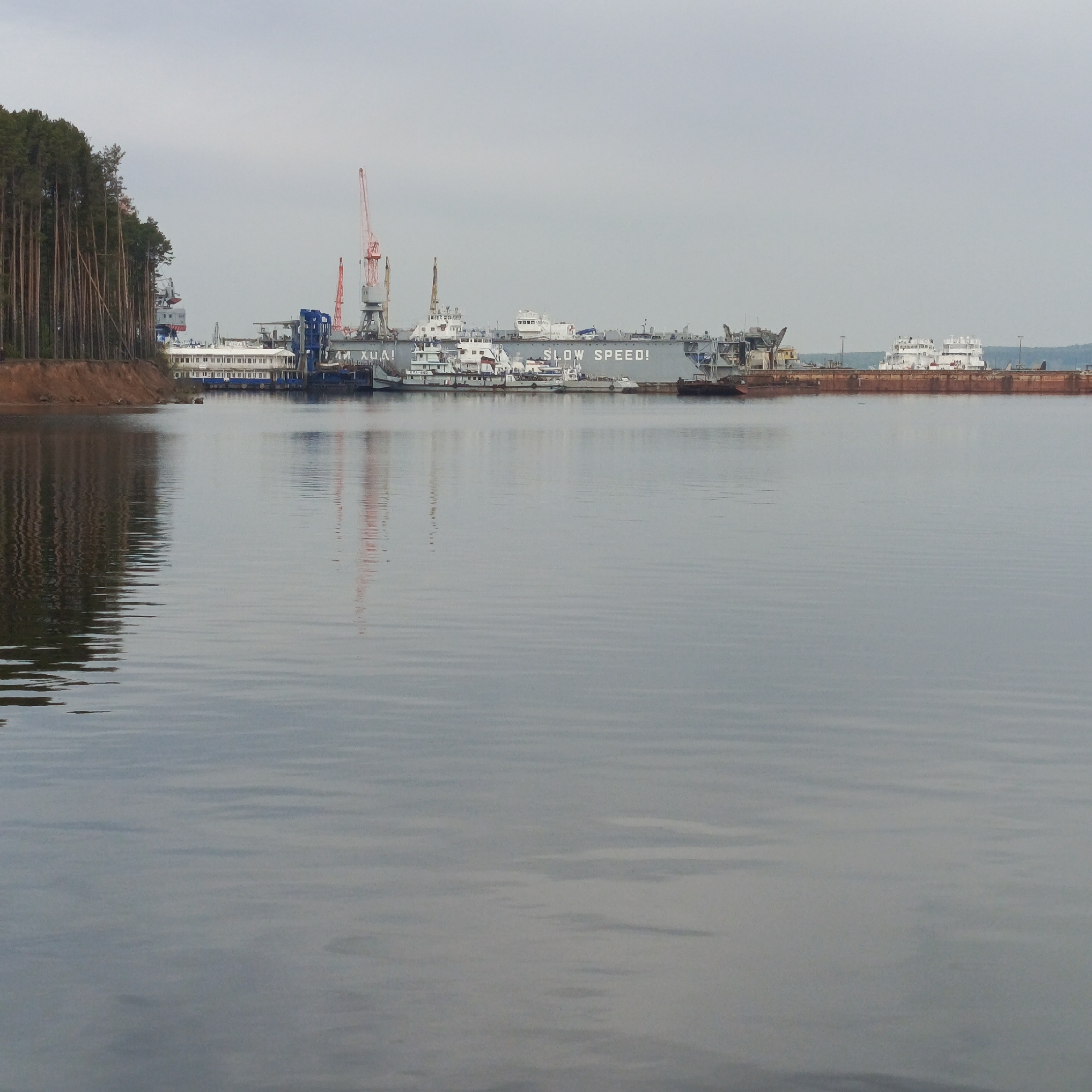 The image depicts a waterfront scene on the Kama River in Chaikovsky, Russia. A shipyard dominates the view, featuring a large floating dock with the text "SLOW SPEED!" visible on its side. Various vessels are docked along the shipyard, including tugboats and what appears to be a passenger ship. A red crane rises above the structures. To the left, a forested shoreline lines the water's edge. The water surface is rippled, and the sky is overcast.