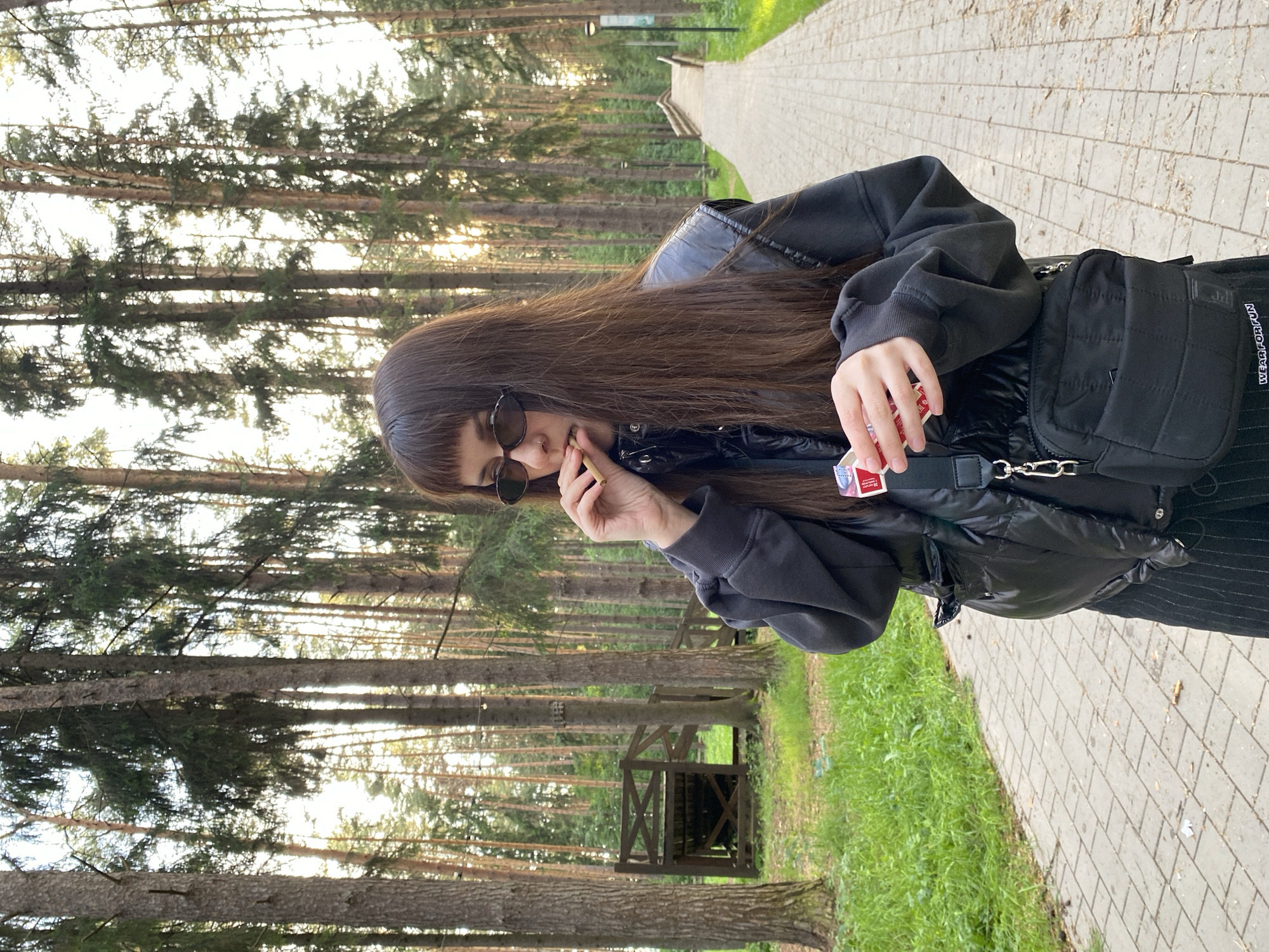 The photograph shows a young woman standing on a paved path in a wooded area. The location is identified as Votkinsk, Russia.
The woman has long, dark brown hair with bangs, and she is wearing sunglasses, a black jacket, and a black bag with a strap across her torso. She is holding a lit cigarette in one hand and a red cigarette box in the other, which she is smoking.
The path extends into the distance, flanked by tall trees on both sides. There is a wooden structure visible among the trees on the left side of the image. The lighting suggests it is either early morning or late afternoon. The overall environment seems like a park or forest area within the city.