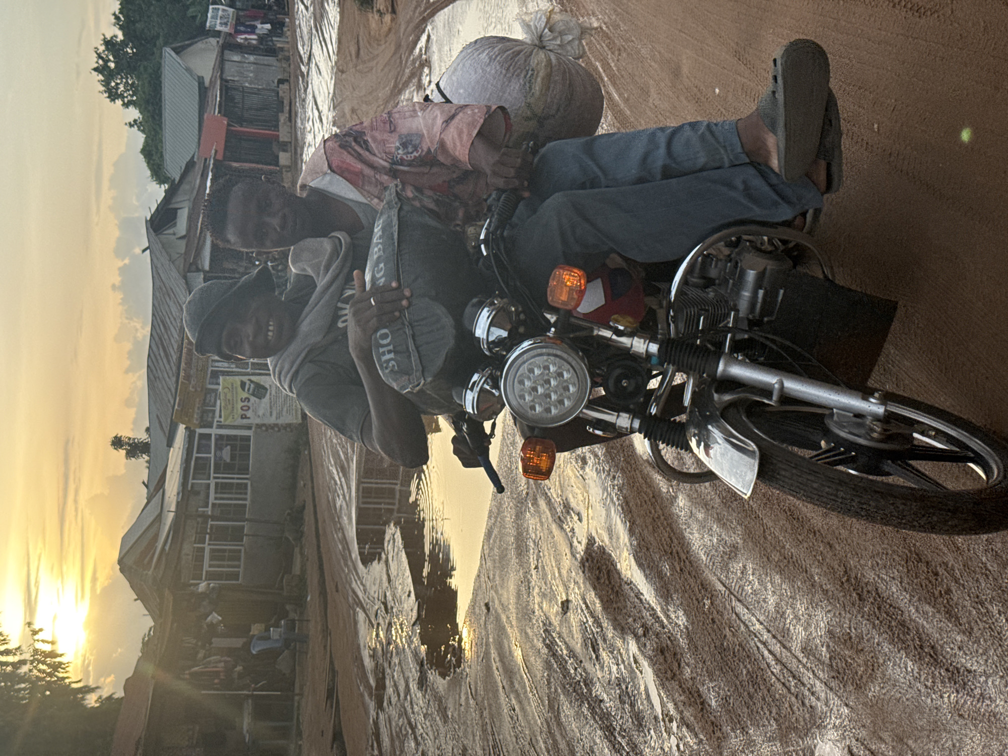 The image depicts two men on a motorcycle on a dirt road in Ihiagwa, Nigeria, under the warm, golden light of either sunrise or sunset. The road is visibly uneven and muddy, with tire tracks and puddles reflecting the low sun.
In the foreground, two men are seated on a utility motorcycle. The man in front wears a grey hooded sweatshirt and a dark cap, smiling towards the camera while holding a small, dark bag with "SHO" (likely part of "SHOPPING BAG") printed on it. The second man, positioned behind the first, wears a patterned long-sleeved shirt and blue jeans, carrying a large, light-colored sack behind him on the motorcycle. His feet, clad in grey sandals, are visible. The motorcycle features a distinctive round LED headlight and orange turn signals.
The background reveals a street scene with various buildings, likely shops or residences, characterized by corrugated metal roofs. A sign with "POS" is faintly visible on one structure. Green trees dot the landscape further back. The overall mood is warm and candid, capturing a moment of everyday life in the community.