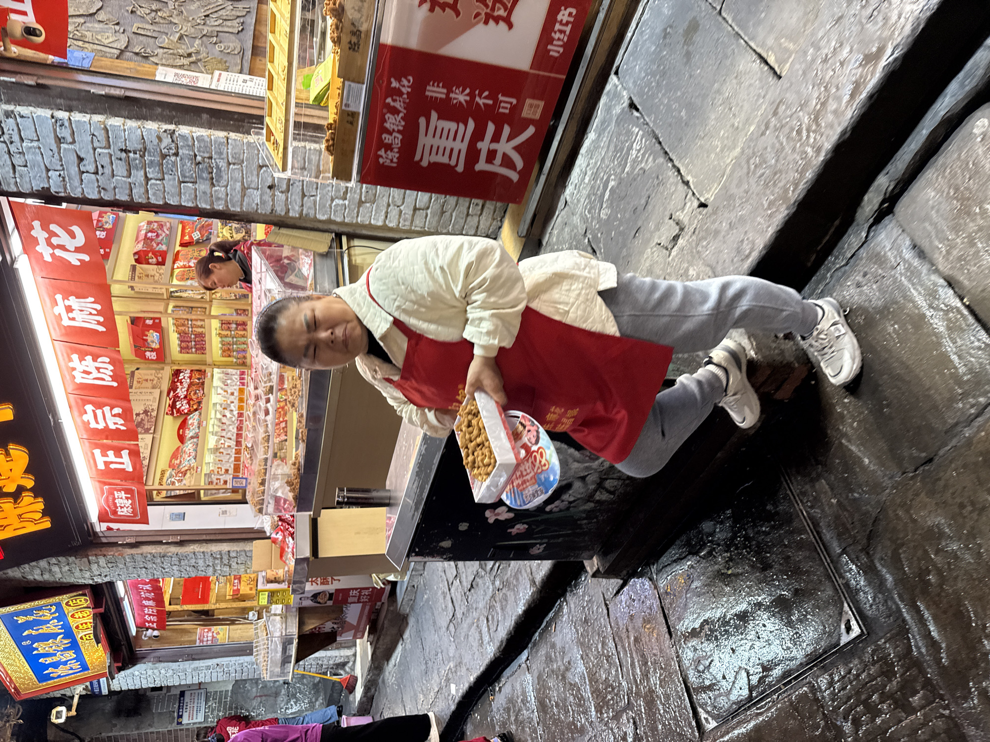 The image shows a vendor in the Shapingba District of Chongqing, China. She is holding a box of twist cookies (ma hua) at a street-side stall. She is wearing a white shirt, grey sweatpants, white sneakers, and a red apron. The stone paved street is damp. The stall advertises "Chongqing twists," and other vendors and shops are visible in the background.