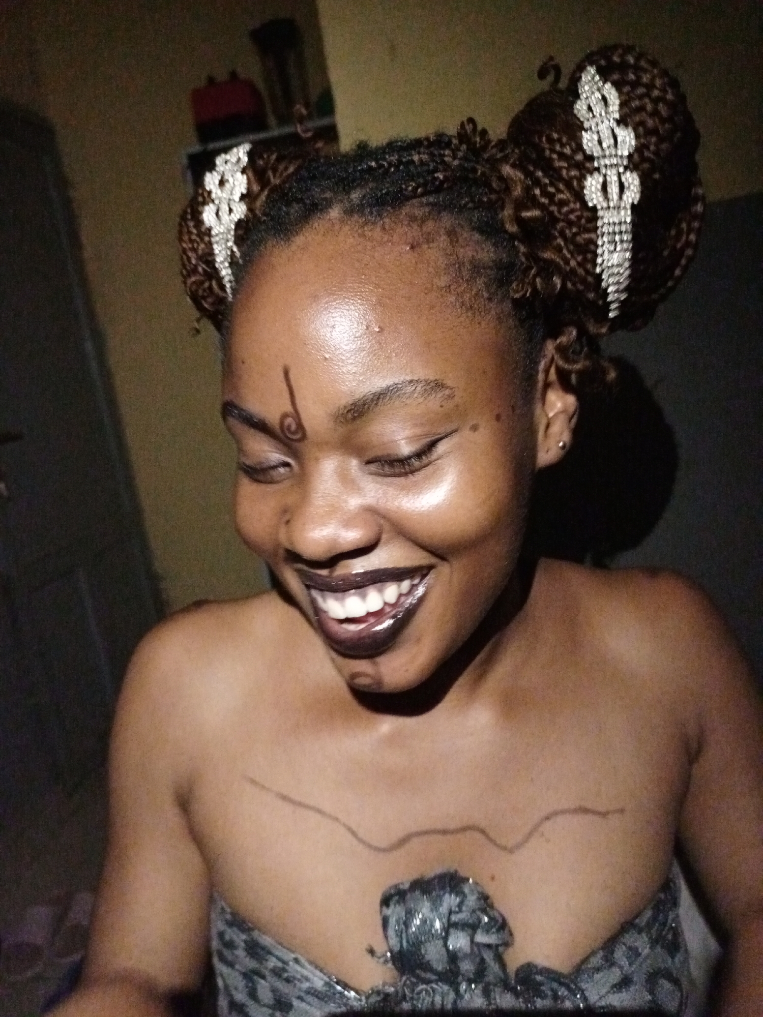 The image captures a young woman from Keffi, Nigeria, smiling broadly with her eyes closed or looking downwards, conveying joy and contentment. Her braided hair is styled into two large buns, each embellished with an intricate, sparkly silver-colored hair accessory. She wears dark, glossy lipstick and features distinctive dark body art: a swirling design on her forehead above her left eyebrow, and a wavy line drawn across her upper chest.
Her skin has a noticeable sheen on her forehead and cheeks, and she wears a small stud earring in her right ear. She is dressed in a strapless garment with a textured, patterned fabric in shades of grey and black, possibly metallic or sequined, visible around her bust. The setting appears to be indoors, dimly lit, with a dark, indistinct background that includes what looks like a shelf with a red object and other items. A direct light source illuminates her face from the front-left, creating highlights and shadows. No text is visible in the image.