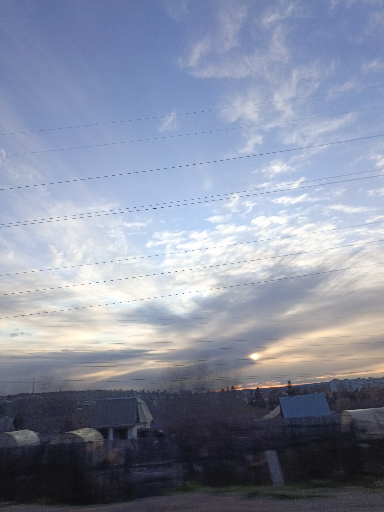 The image depicts a landscape at dusk, possibly in the vicinity of Chaikovsky, Russia, given the urban and rural characteristics. A series of high-voltage power lines span across the sky, which is characterized by a mix of blue hues and various cloud formations. The lower portion of the sky exhibits a warmer, sunset-like glow. Below the horizon, a residential area is visible. Buildings with sloped roofs are apparent, partially obscured by trees and shrubs. The foreground shows vegetation suggestive of a roadside or field. There are no individuals visible in the image. The overall atmosphere is one of quiet tranquility.