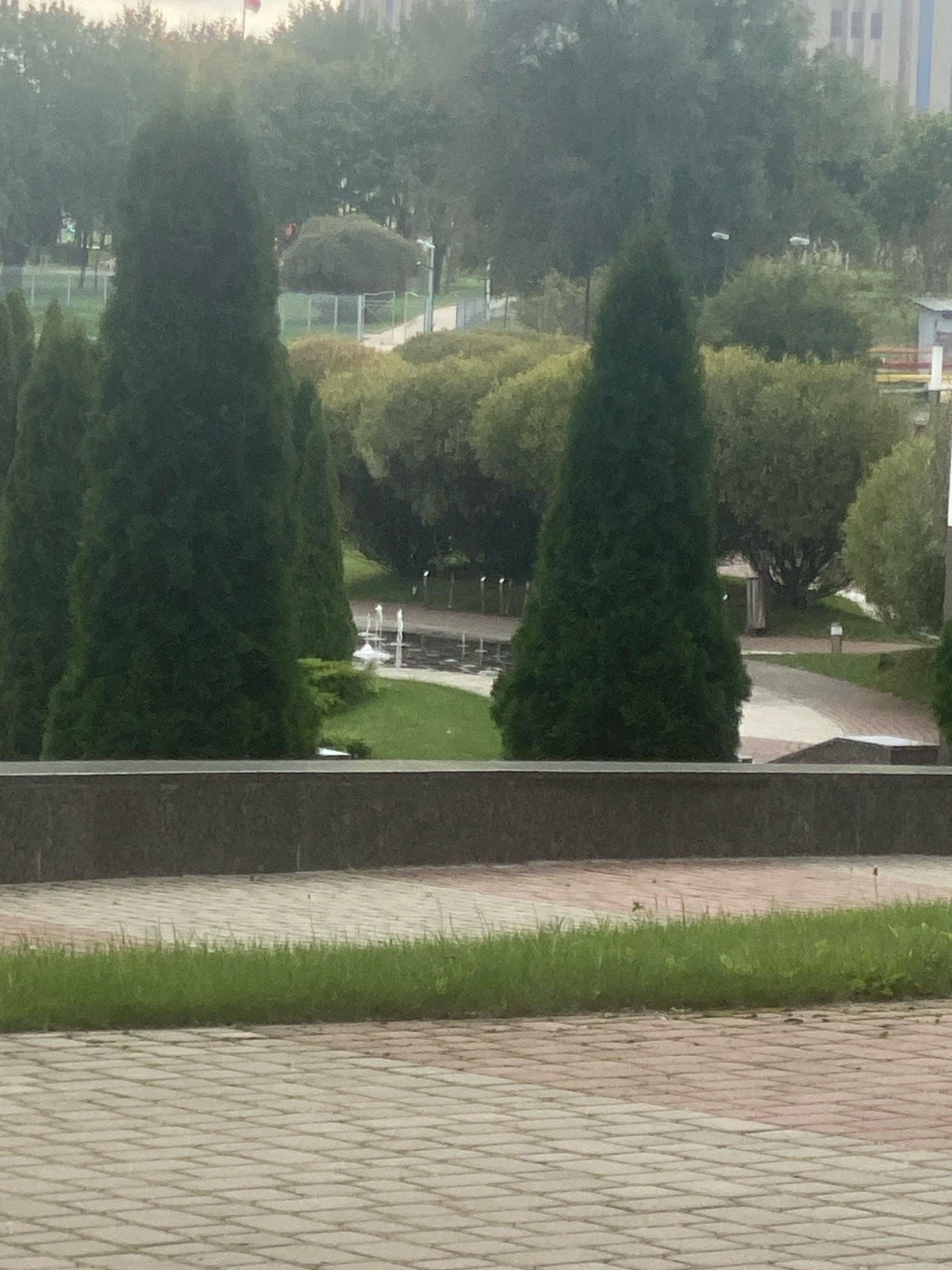 The image depicts a public park area in Minsk, Belarus, characterized by mature trees, manicured bushes, and paved walkways. In the foreground, a wide, stone-bordered pathway leads to a grassy area. Beyond this, a pathway curves through a park with several tall, conical evergreen trees and shorter, rounded bushes. A fountain is visible in the mid-ground, with water jets arcing upwards from multiple points within a circular pool. In the background, the park continues with more trees, some appearing to be deciduous, and buildings can be seen in the distance, suggesting an urban setting. The sky is overcast, giving the scene a muted, natural light. There are no people visible in the image.