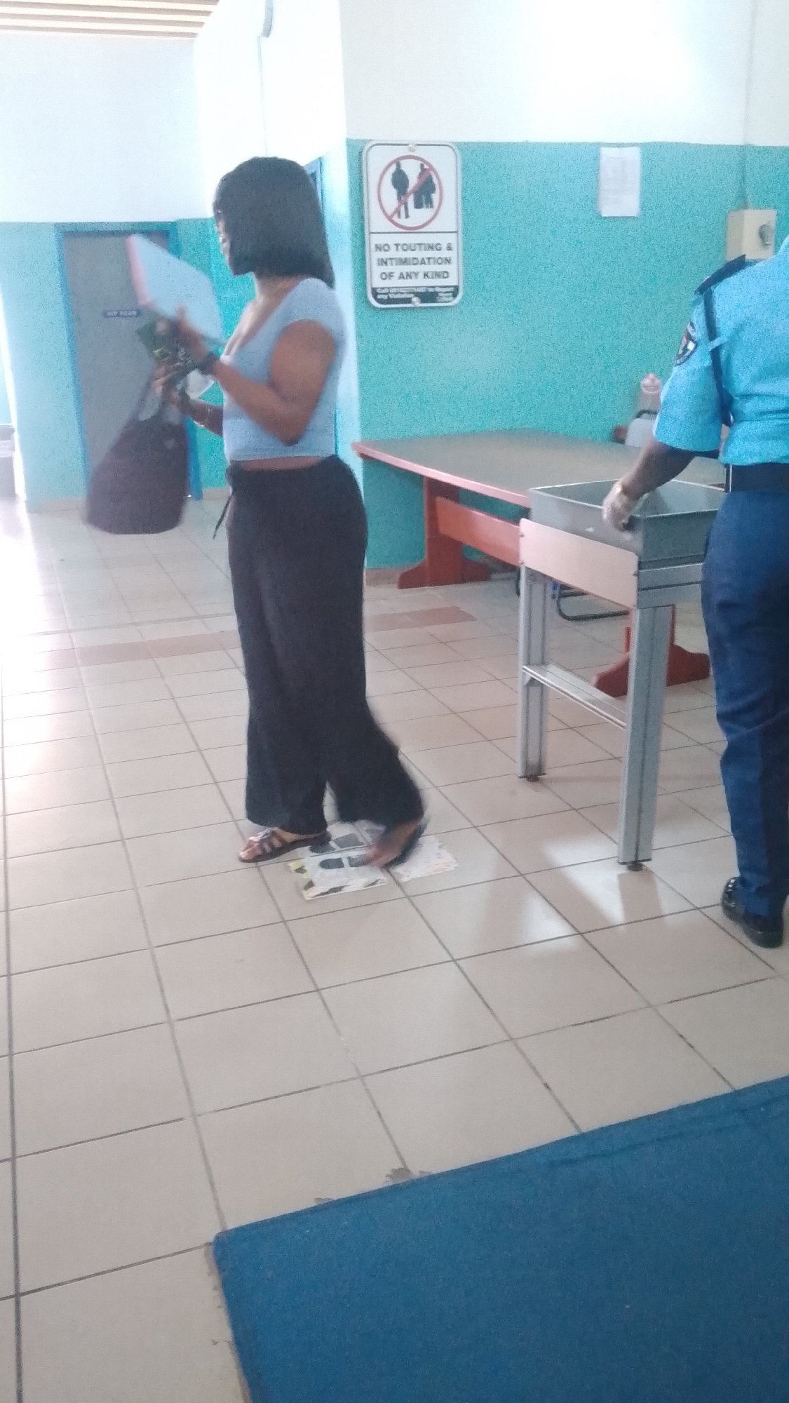 Inside a building in Orerokpe, Nigeria, a woman with short black hair is passing through a security checkpoint. She wears a light blue shirt and black pants, holding a brown bag. She stands on a patterned floor tile marked with shoe placement guides. A uniformed security officer stands at the metal detector table, wearing white gloves. A sign on the wall reads "NO TOUTING & INTIMIDATION OF ANY KIND."