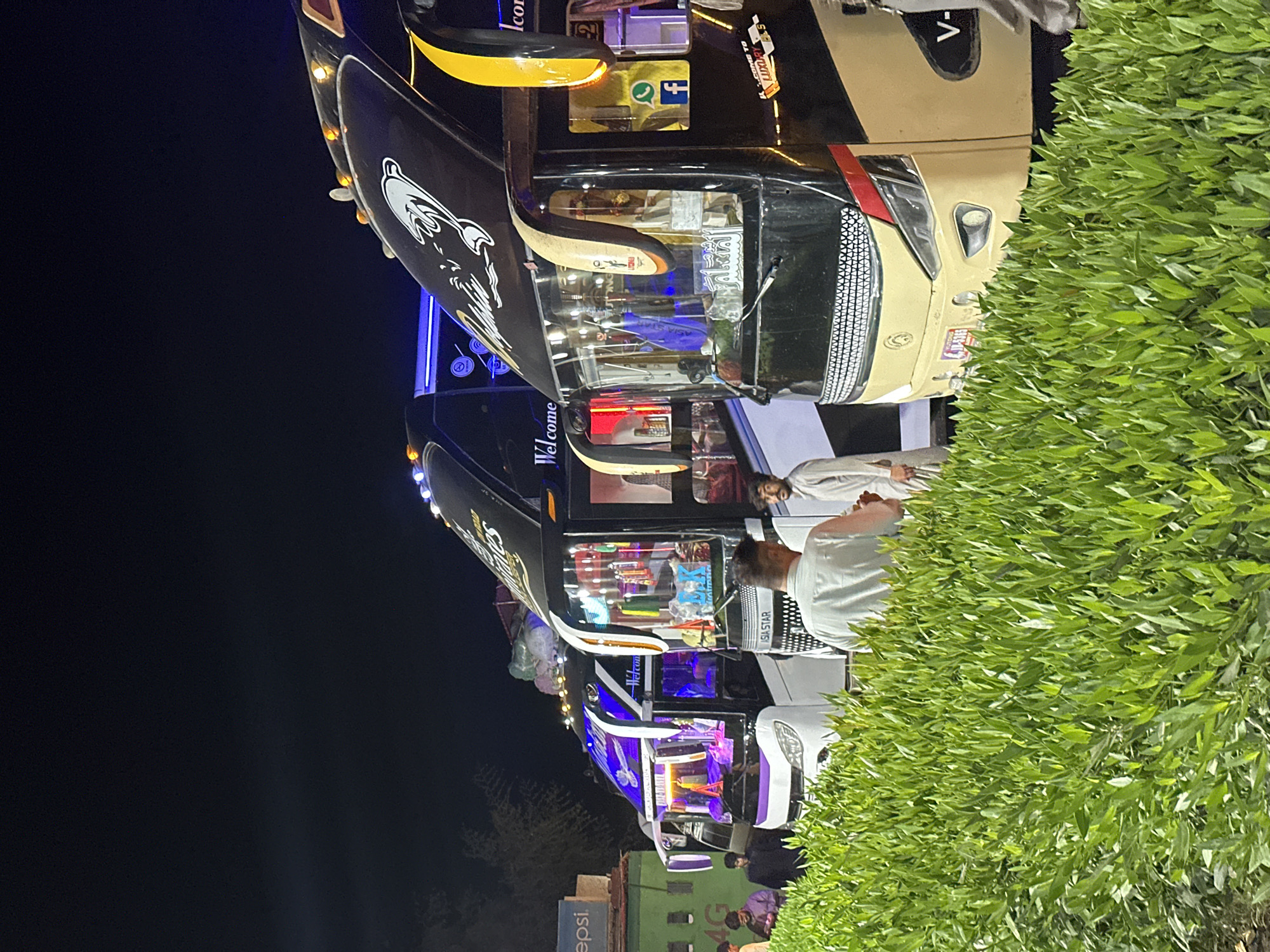The image captures a nighttime scene featuring several brightly lit and highly customized vehicles, resembling buses or large vans, parked outdoors. A dense, verdant green hedge runs along the right side of the frame, partially obscuring the view of the vehicles and people.
The main subjects are the vehicles and a few individuals standing nearby. The vehicles are elaborately decorated with numerous lights, intricate designs, and often display goods or interiors illuminated with various colors, suggesting they might function as mobile shops, food stalls, or specialized passenger transport. One prominent vehicle is black and tan/cream colored, featuring a detailed grille and what appears to be a graphic of a dolphin or fish. Another black vehicle behind it has "Welcome" written on its side. People are observed casually standing around these vehicles, some appearing to interact with what's inside.
The time of day is clearly night, evidenced by the dark sky and the reliance on artificial lighting from the vehicles. The setting appears to be an outdoor area, possibly a roadside stop or an informal gathering place.
Visible text includes "epsi" (part of a Pepsi logo) and "4G" on a distant sign. On the vehicles or associated items, "ASIA STAR," "Welcome," "المسلم" (Arabic text), "LUXURY," "Icon," and "V-" are visible. Social media logos for Facebook and WhatsApp are also present, alongside the word "COME TO" which is partially visible.