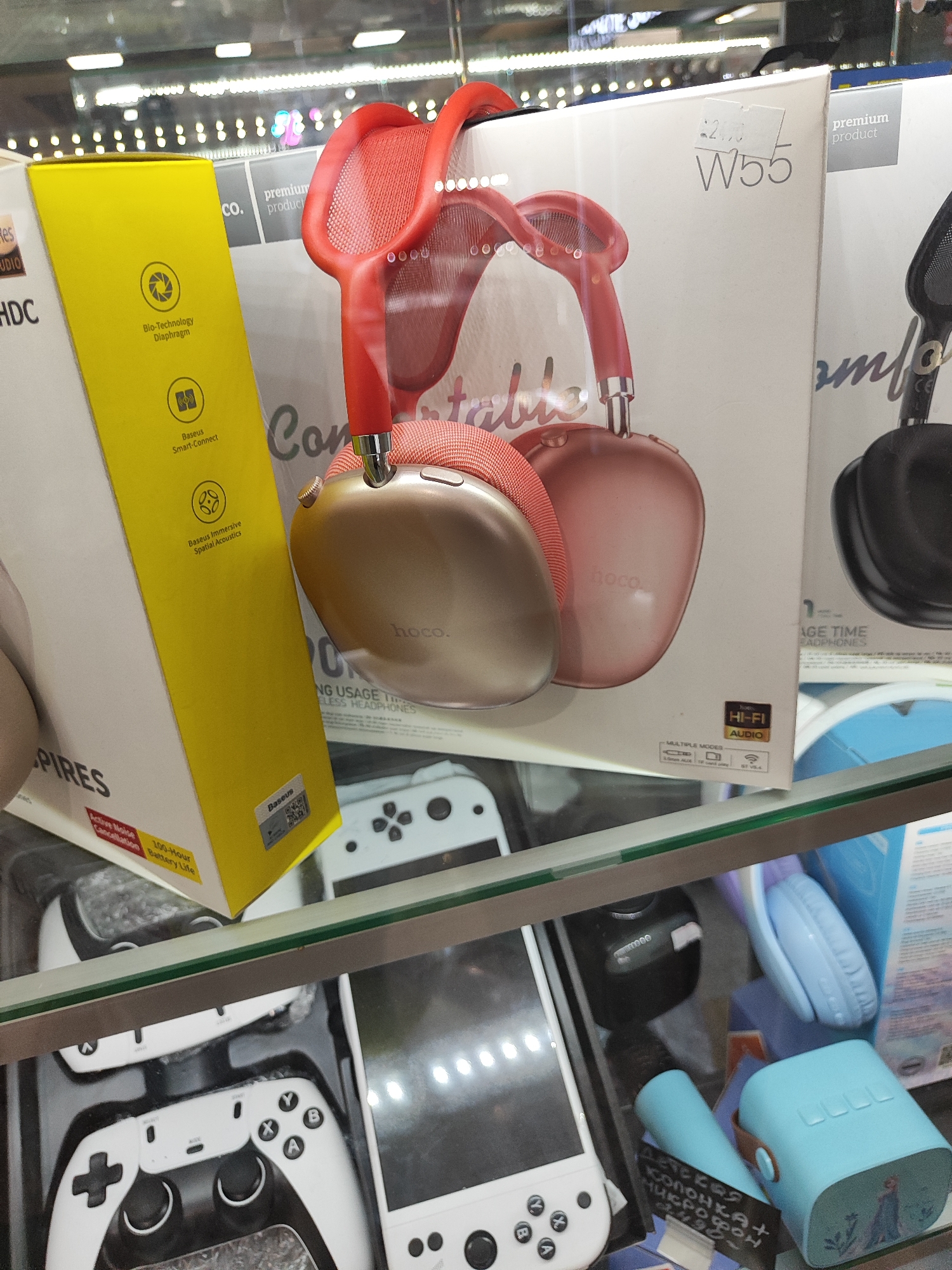 The image shows a display of electronics in a store. In the foreground, a clear glass shelf exhibits various electronic devices. On the left side of the shelf, a yellow box with text and icons is visible, along with a gold and red pair of headphones on top of it. To the right of the headphones, a white box with the model "W55" and the text "premium product" is displayed. Further to the right, a black pair of headphones is partially visible. Below the primary display, another shelf holds white game controllers and a white handheld gaming device. On the lower right side of the image, a blue portable speaker with a design of characters and a blue headset are present. The lighting suggests an indoor retail environment. The city and specific location are unknown.