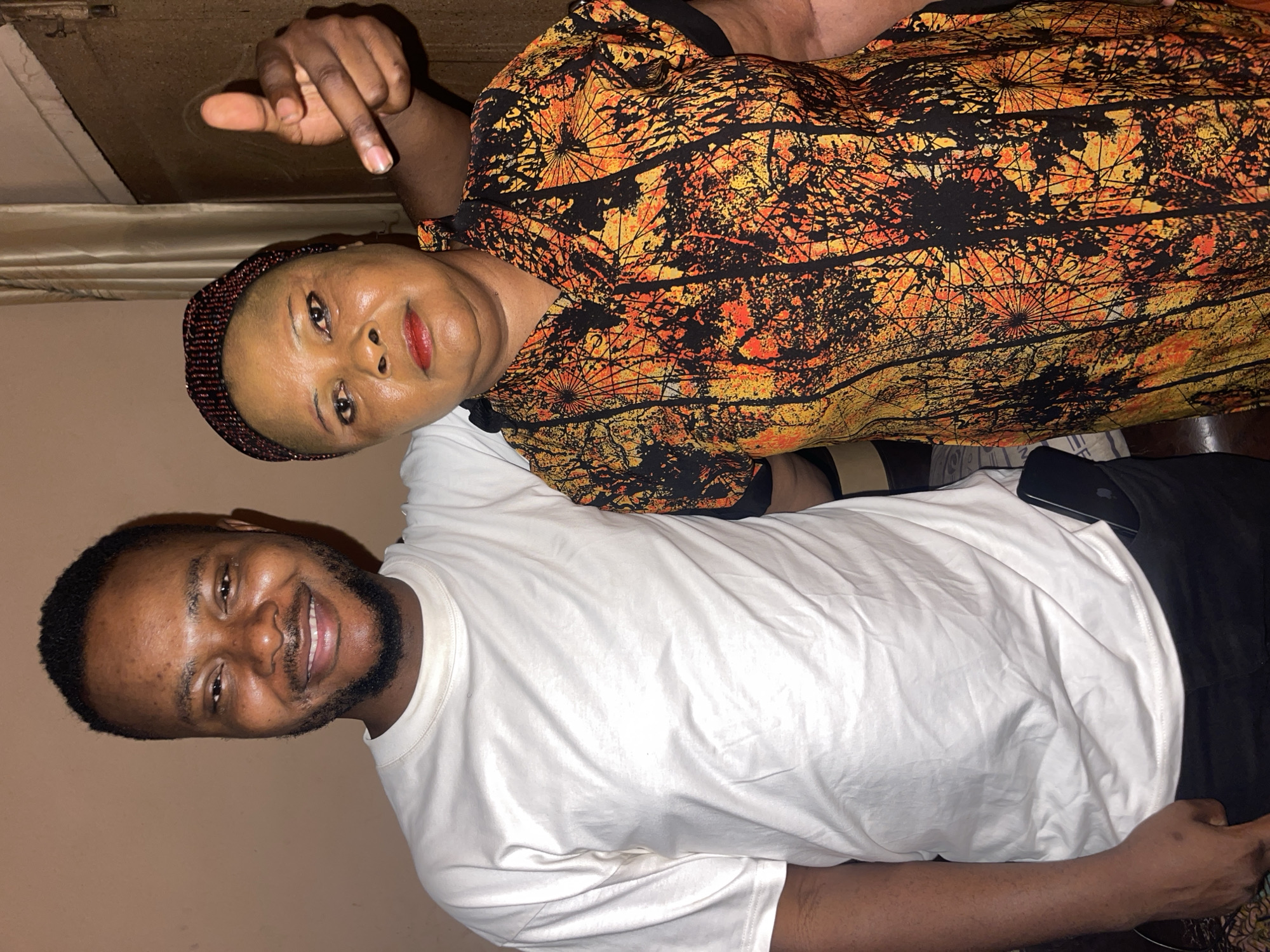 Two individuals are posing for a photograph. A woman with a dark complexion, wearing a head covering and a brightly colored patterned top, is positioned at an angle to the camera, pointing a finger towards the top of the frame. A man with a white t-shirt and a beard is standing next to her, smiling at the camera. The background is a plain wall. It is not possible to infer the location of Muchiya, Nigeria, from the image.