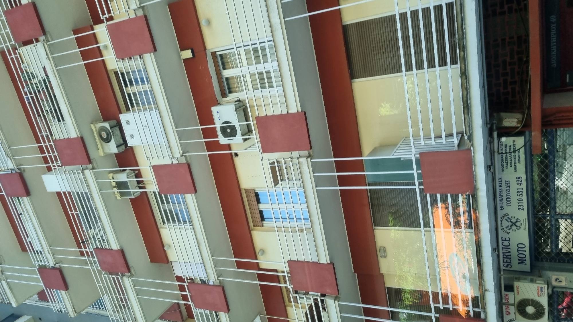 The image shows a multi-story building with balconies and air conditioning units. On the ground floor, there is a sign for "SERVICE MOTO" which appears to be a motorcycle repair shop. The sign also includes a phone number and text in Greek, indicating services such as engine repair and parts. The building is painted in light yellow and has red accents on the balconies. Some windows have blinds. The overall impression is of an urban environment, likely in Greece, given the Greek text on the sign.