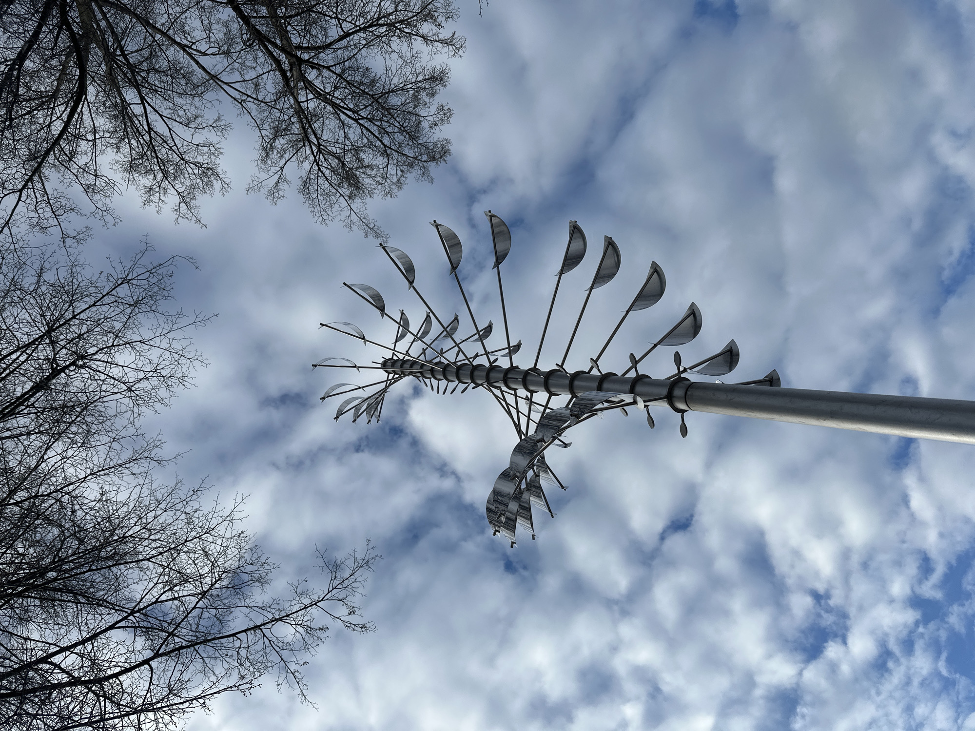 The image shows a low-angle view of a kinetic sculpture against a backdrop of a cloudy, blue sky. The sculpture is mounted on a gray, cylindrical pole. It consists of multiple horizontal rods branching out from the main pole, each adorned with several small, curved, metallic 'leaves' or 'vanes'. The structure has a symmetrical, slightly top-heavy appearance. Bare tree branches are visible in the top left and bottom left corners of the frame, obscuring the view. The setting is Votkinsk, Russia, implied by the presence of this distinctive local sculpture.