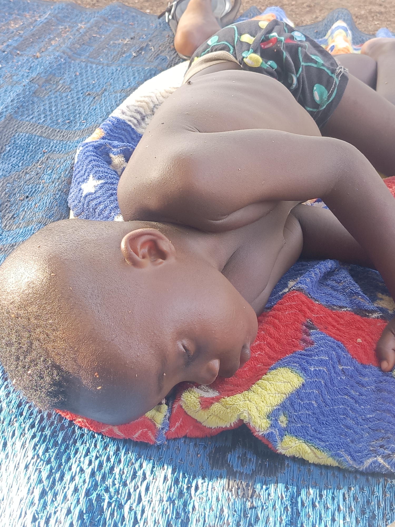 A child is sleeping on a colorful blanket in Akwanga, Nigeria. The child is lying on their side, with their head resting on the blanket. The child is wearing shorts. The blanket is on top of a blue plastic mat. In the top left corner is a sandal.
