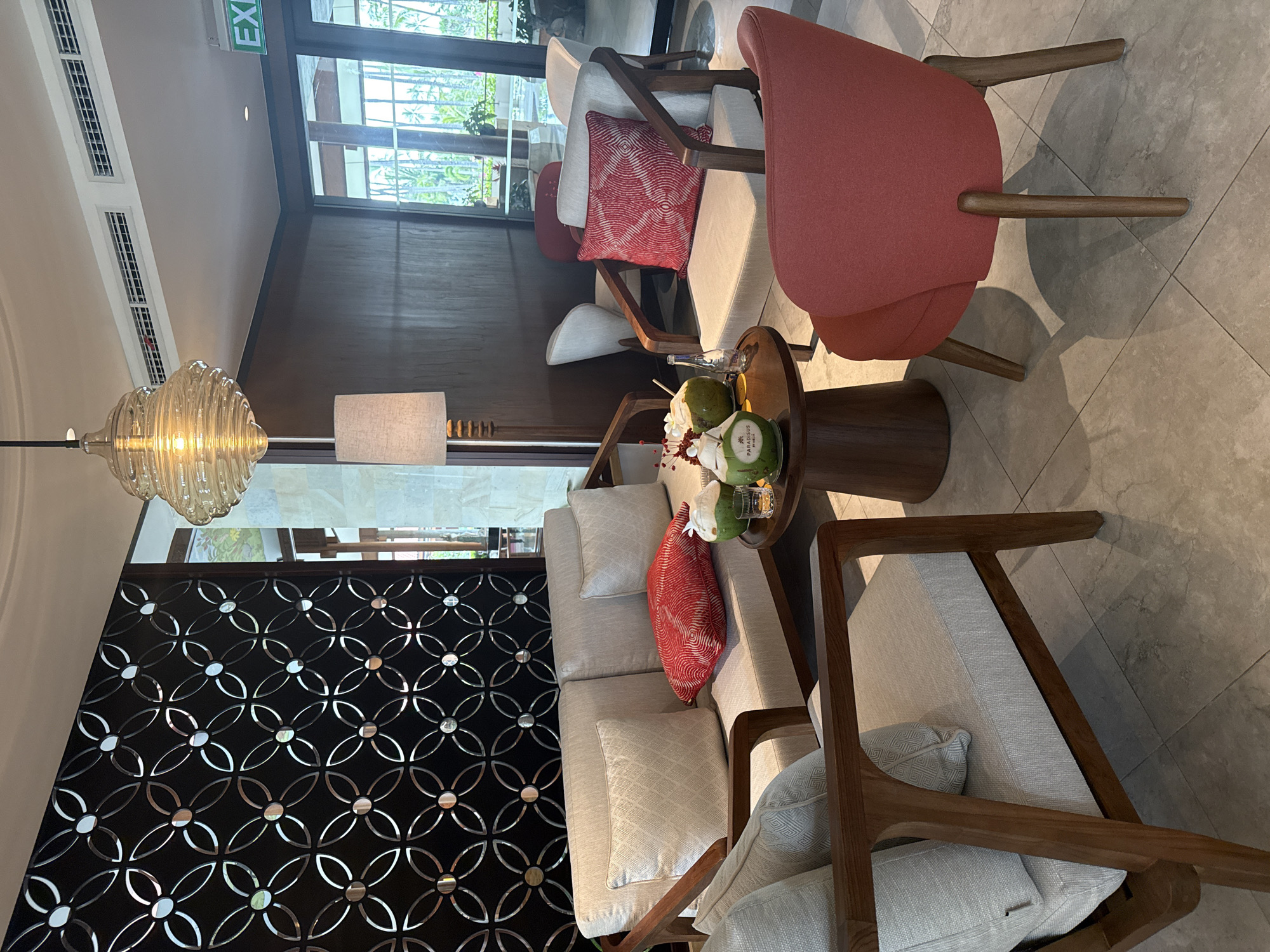 This image shows a seating area in a hotel lobby in Kuta Selatan, Indonesia. The scene features a neutral-colored sofa and two armchairs, all with wooden frames and light-colored cushions. A red patterned cushion rests on the sofa, and another similar red cushion is on one of the armchairs. On a small, round wooden coffee table, there are two whole coconuts with straws, a small glass with a golden liquid, and a few decorative red elements. Behind the seating area, a decorative black screen with a repeating circular pattern is visible. A floor lamp with a beige shade stands to the left of the sofa. In the background, large glass doors open to an outdoor view with palm trees. An "EXIT" sign is visible above the doors. The flooring is tiled with a light gray, stone-like pattern. The overall ambiance is relaxed and tropical.