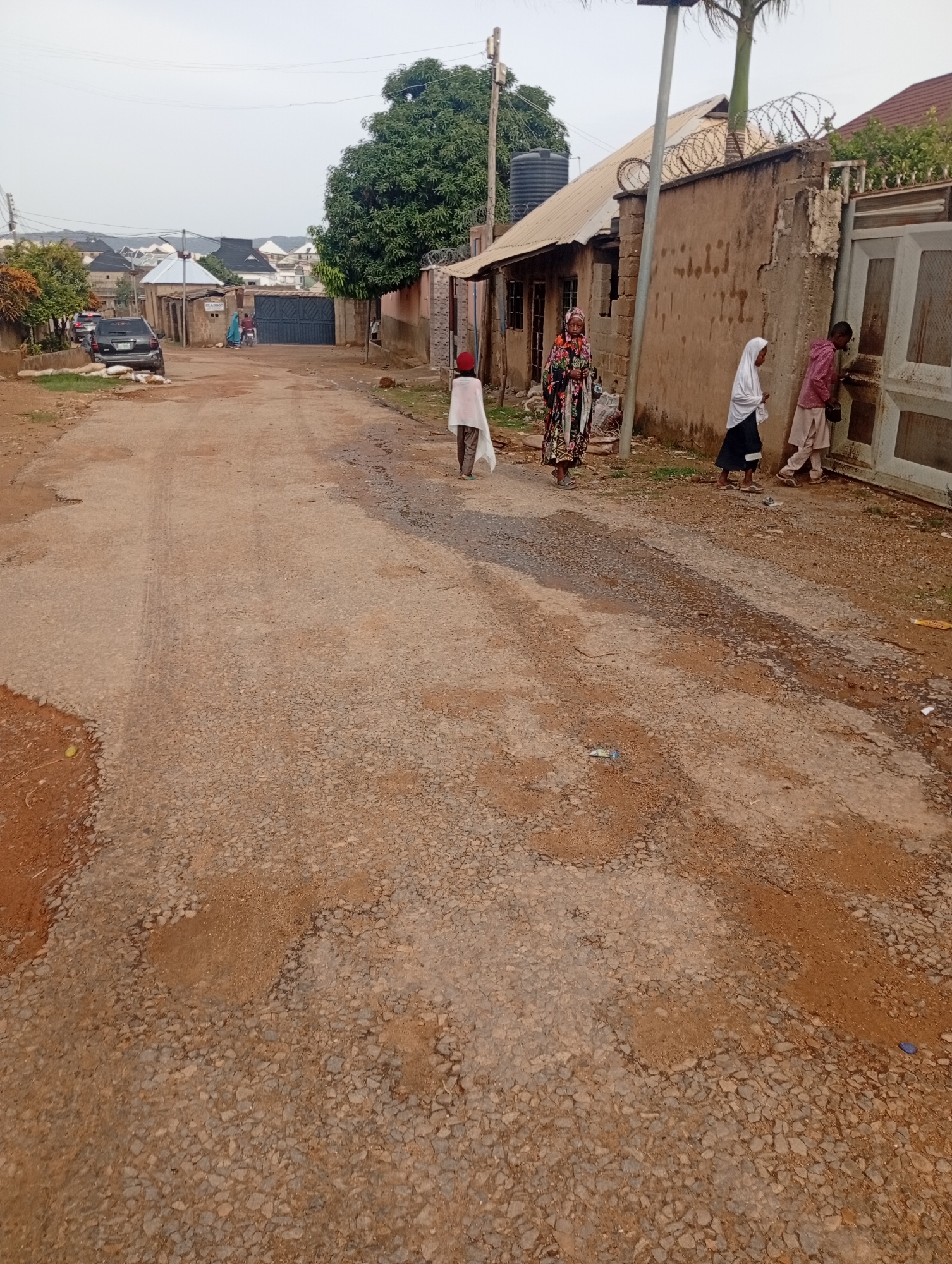 The image captures a street scene in the city of Rafin Par, Nigeria. The road is unpaved and appears to be in a state of disrepair, with gravel and patches of dirt. Several people are present: a woman in a colorful dress, a child in a white garment and red head covering, a young woman in a white hijab and black skirt, and a boy in a pink shirt and beige trousers. The boy is interacting with a gate on the right side of the image. In the background, there are buildings, some with corrugated metal roofs, and a large tree. A black SUV is parked on the left side of the road. The sky is overcast, suggesting cloudy weather.