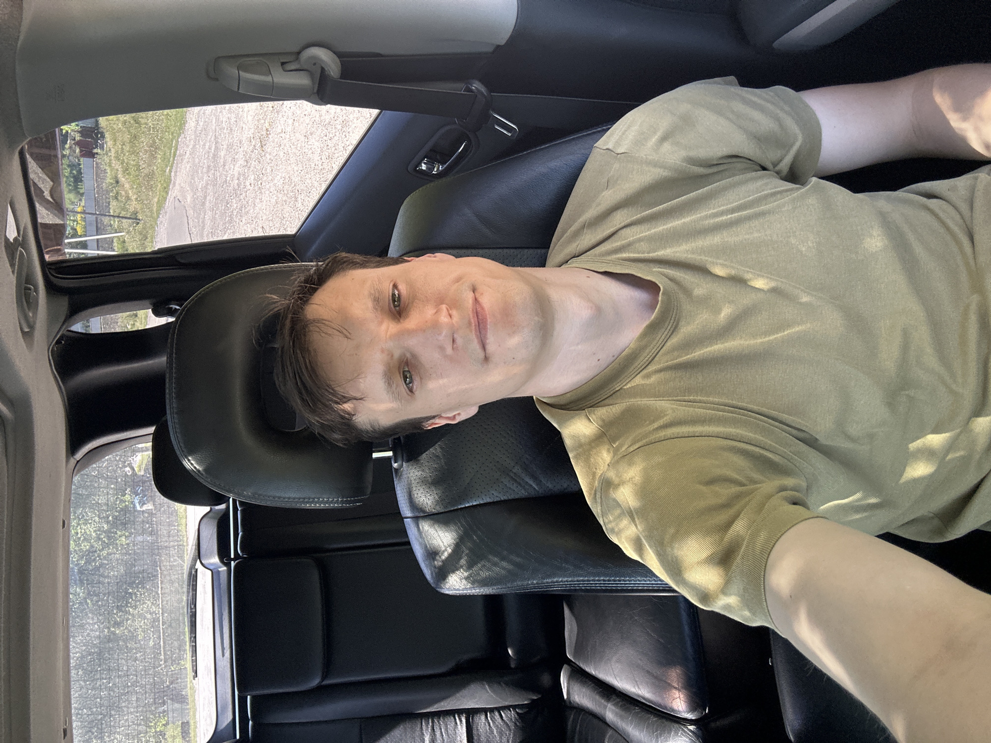 A man with short brown hair and green eyes sits in the driver's seat of a car, looking directly at the camera with a slight smile. He is wearing a light olive green t-shirt. The interior of the car features black leather seats and a sunroof. Through the windows, a rural outdoor scene is visible, with trees and a dirt road. This image was taken in the village of Makiv, Ukraine.