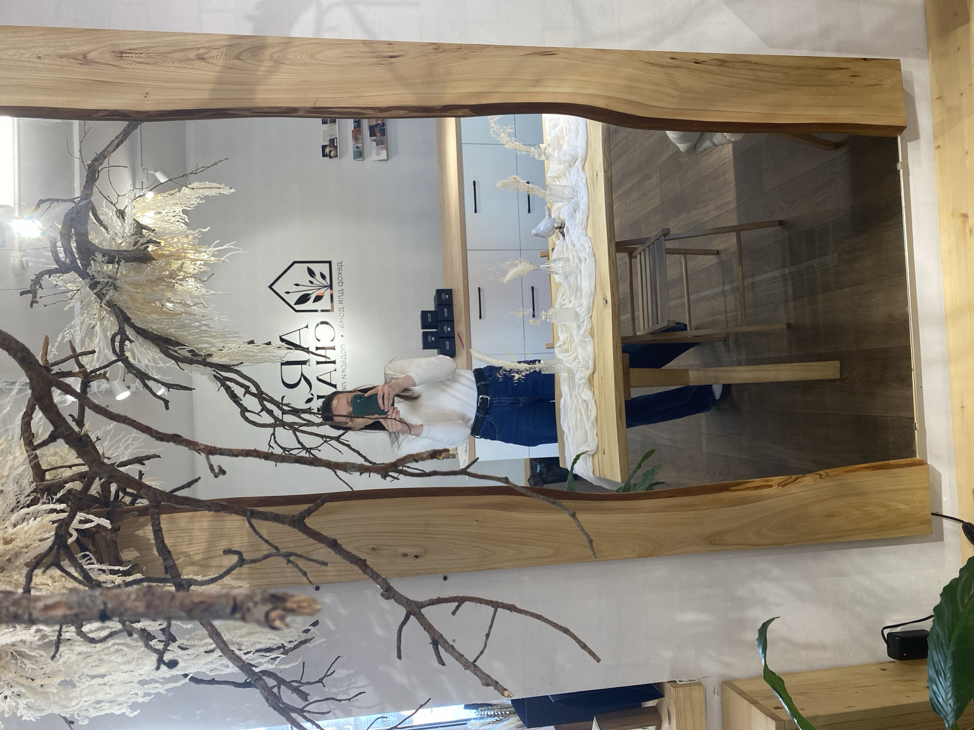 The image depicts a woman standing in front of a large mirror with a natural wood frame. She is holding a smartphone and appears to be taking a picture of herself in the mirror. Her reflection shows a white long-sleeve shirt and blue jeans.
The mirror reflects a shop interior. On the left side of the mirror, there is a wall with a company logo that reads "АЙ ДА САД", along with some text in Russian. Below are several cosmetic products or packaging. To the left of the logo is an elaborate dry floral and branch display.
In the background, there is a white cabinet with drawer pulls. In front of the cabinet is a dining table set up with a white runner and vases filled with dried flowers. A wooden chair is situated next to the table. The floor appears to be wooden.
The background includes interior elements, such as the white cabinet, table setting, and general shop decor. A power outlet is also visible behind the mirror.
Based on the logo and text displayed in the image, the location is likely in Chaikovsky, Russia, as it corresponds with the Russian text accompanying the logo.
