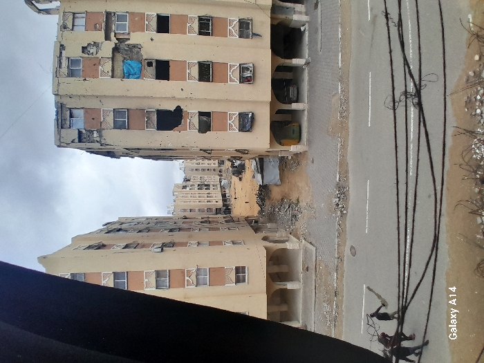 The image depicts a devastated urban landscape in Khan Yunis, Palestinian Territory, under an overcast sky. Two heavily damaged, multi-story residential buildings, constructed with light brown facades and decorative white window surrounds, flank a rubble-strewn alleyway.
The building on the left exhibits numerous pockmarks, large sections of missing facade revealing internal structures, and shattered or absent windows across multiple floors. Its top edge is visibly crumbled. The ground floor features arched open bays.
The building on the right presents similar extensive damage, including large holes in its facade and missing windows, some of which are covered by black tarps or fabric. A notable blue tarp covers a section on the fourth floor from the bottom. The top portion of this building also shows severe structural damage. Its ground floor contains arched openings, with a yellow vehicle and a silver vehicle visible parked within.
Between these two primary structures, a narrow, unpaved path or alley is filled with debris, concrete rubble, and twisted metal. A small, makeshift shelter or structure with a corrugated roof is situated in this debris-laden passage. Further down the alley, additional damaged buildings recede into the distance, and faint figures are discernible walking through the rubble.
In the foreground, an asphalt road with double white center lines and a manhole cover is visible. Several black cables run horizontally across the mid-foreground. Three individuals are on the road in the left foreground, walking towards the left. One person in dark clothing carries a long, light-colored object, possibly fabric or a blanket, over their shoulder. Below the road, tangled wires or rebar are visible. The image bears a "Galaxy A14" watermark in the bottom left corner.