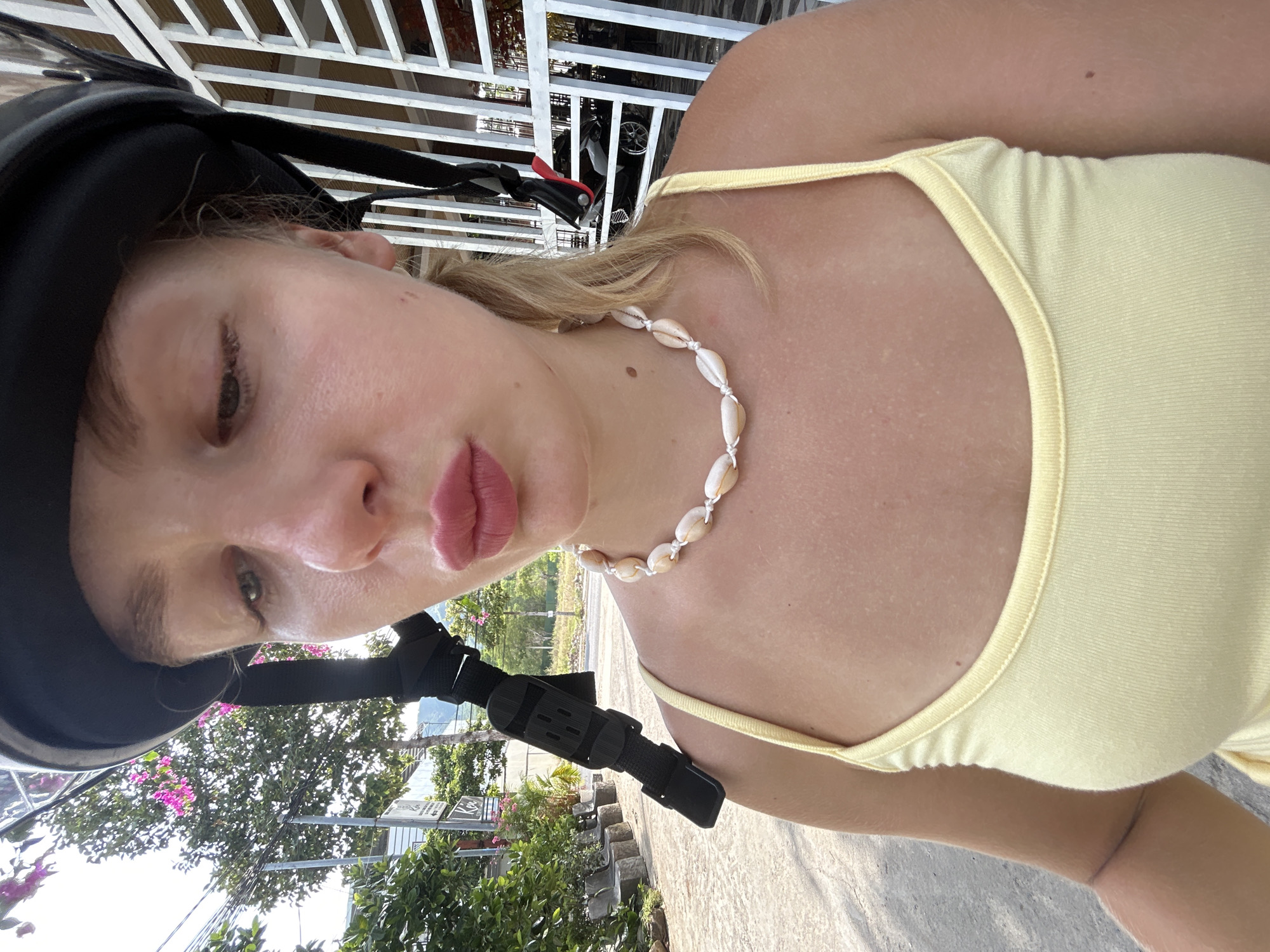 This is a low-angle, close-up selfie of a young woman wearing a black helmet and a pale yellow tank top. She is wearing a necklace made of small white shells. Her blonde hair is visible around the helmet. Her eyes are open and looking towards the camera, and her lips are slightly pursed. The photo appears to be taken outdoors during the day, with sunlight illuminating her face and chest. In the background, there is lush green foliage, trees with pink flowers, and a white railing structure. There is also a black strap with plastic buckles visible on the left side of the frame, which may be part of the helmet or a camera mount. The scene suggests a warm, tropical environment. The overall mood is casual and relaxed. There are no discernible signs or text visible in the image.