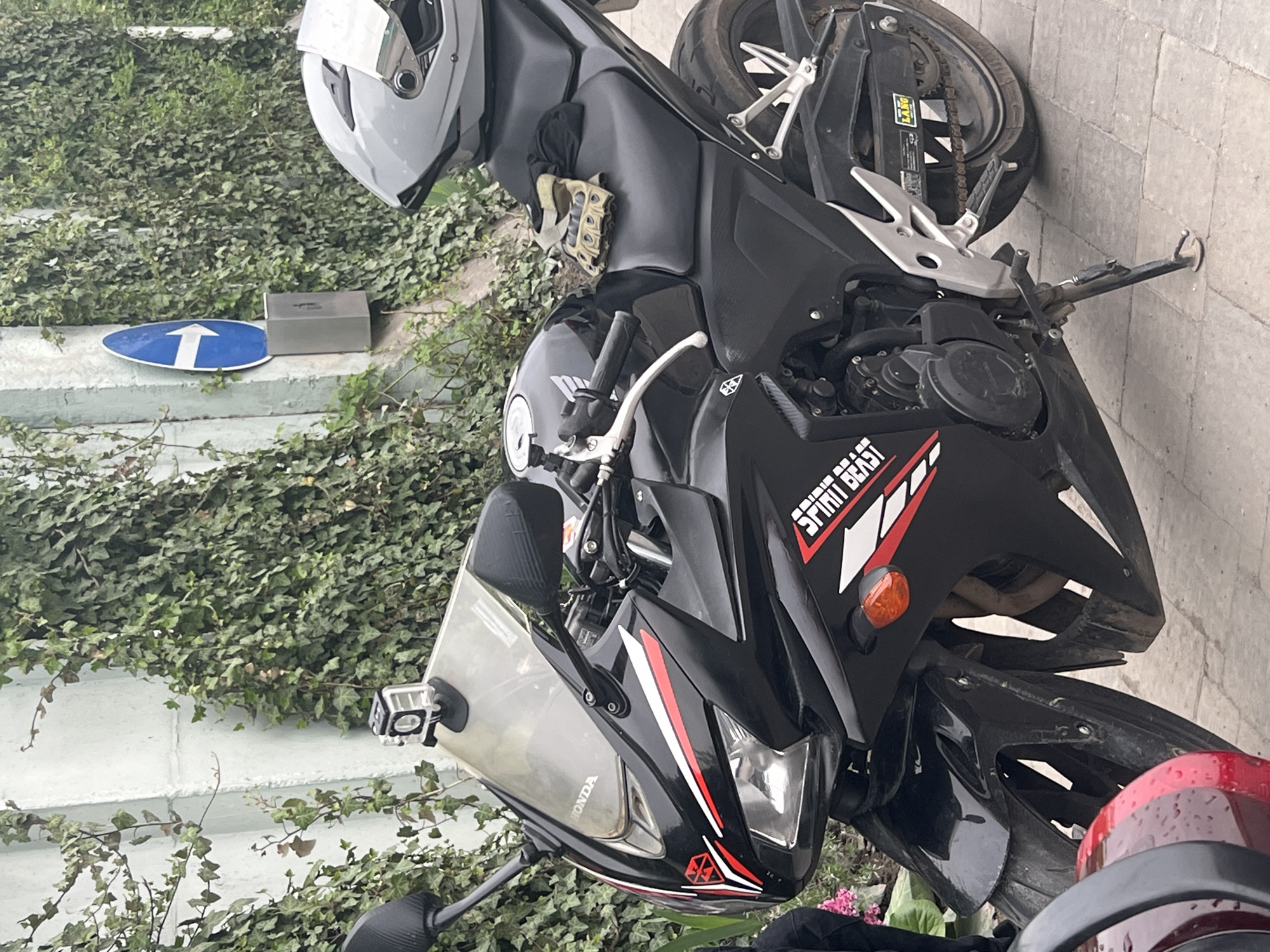A motorcycle is parked on a paved surface. The motorcycle is a black Honda with red and white accents, and the words "Spirit Beast" are visible on the side. A grey helmet rests on the bike's seat. Gloves are on the bike in front of the helmet. The background features a wall covered in ivy and a blue sign with a white arrow. The general location could be Horodok, Ukraine, as requested.