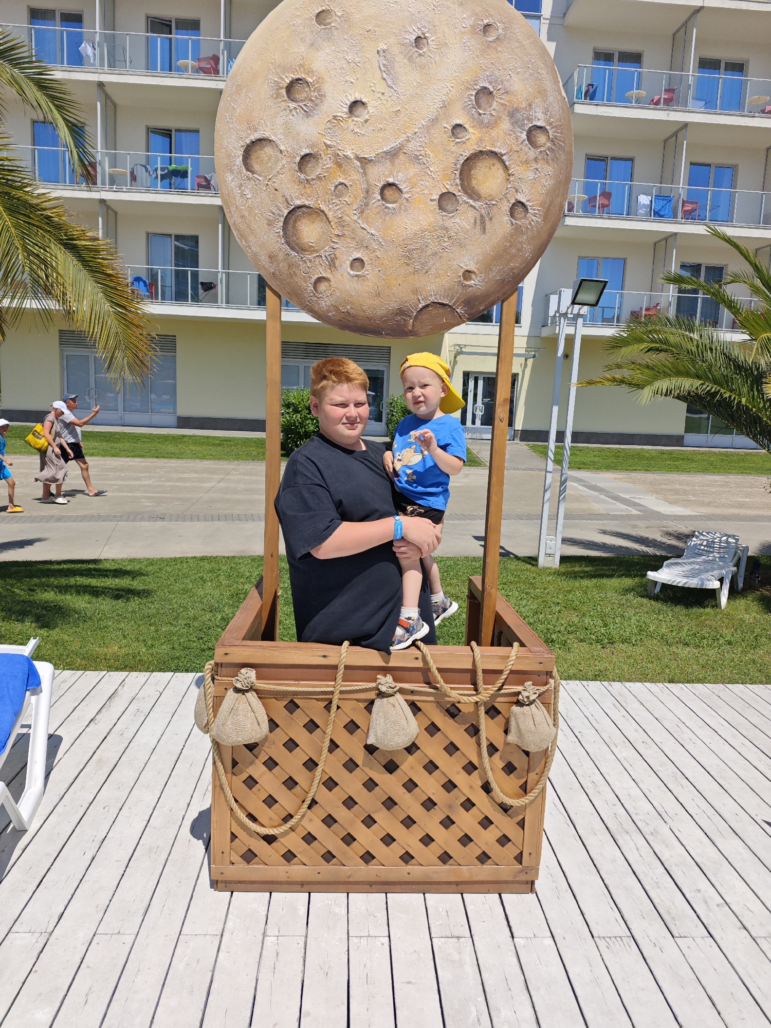 Two young boys are posing in a decorative hot air balloon basket, which is part of a prop that resembles the moon, situated outdoors on a wooden deck. The older boy, with red hair and wearing a black t-shirt, holds the younger boy in his arms. The younger boy, wearing a yellow cap and a blue t-shirt with a cartoon character, looks towards the camera. In the background, a multi-story hotel with balconies is visible, along with palm trees and a lawn. The overall setting suggests a resort or recreational area, potentially in Sochi, Russia, given the presence of palm trees and the style of the hotel, which is common in Black Sea coastal resorts. The sun is shining, casting shadows, indicating it is daytime.