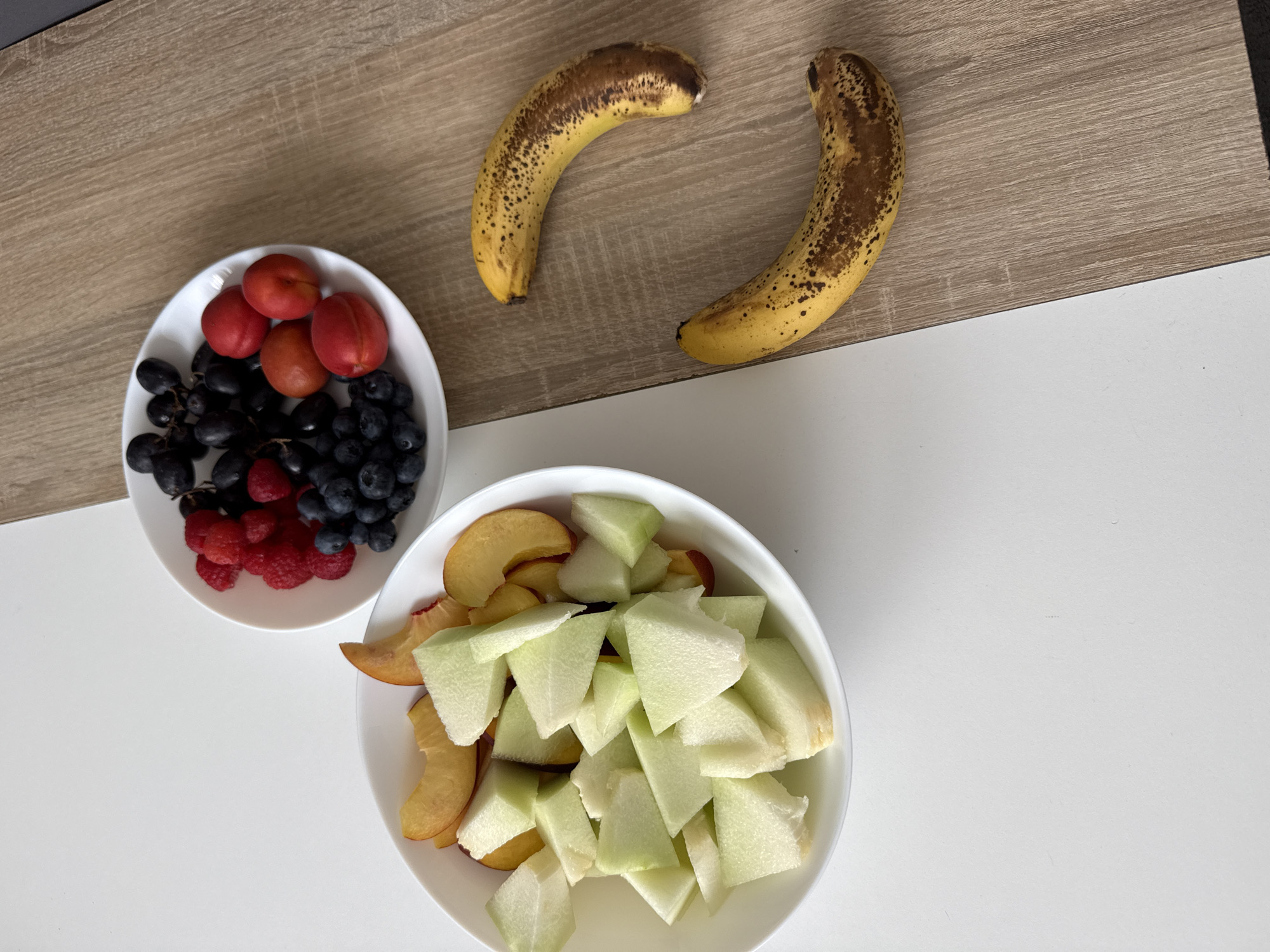 In Hanau, Germany, a still life composition features a white bowl filled with chopped honeydew melon and sliced peaches. To the right, a white plate holds dark grapes, raspberries, blueberries, and red plums. Two speckled bananas lie on a wooden surface adjacent to the white background.