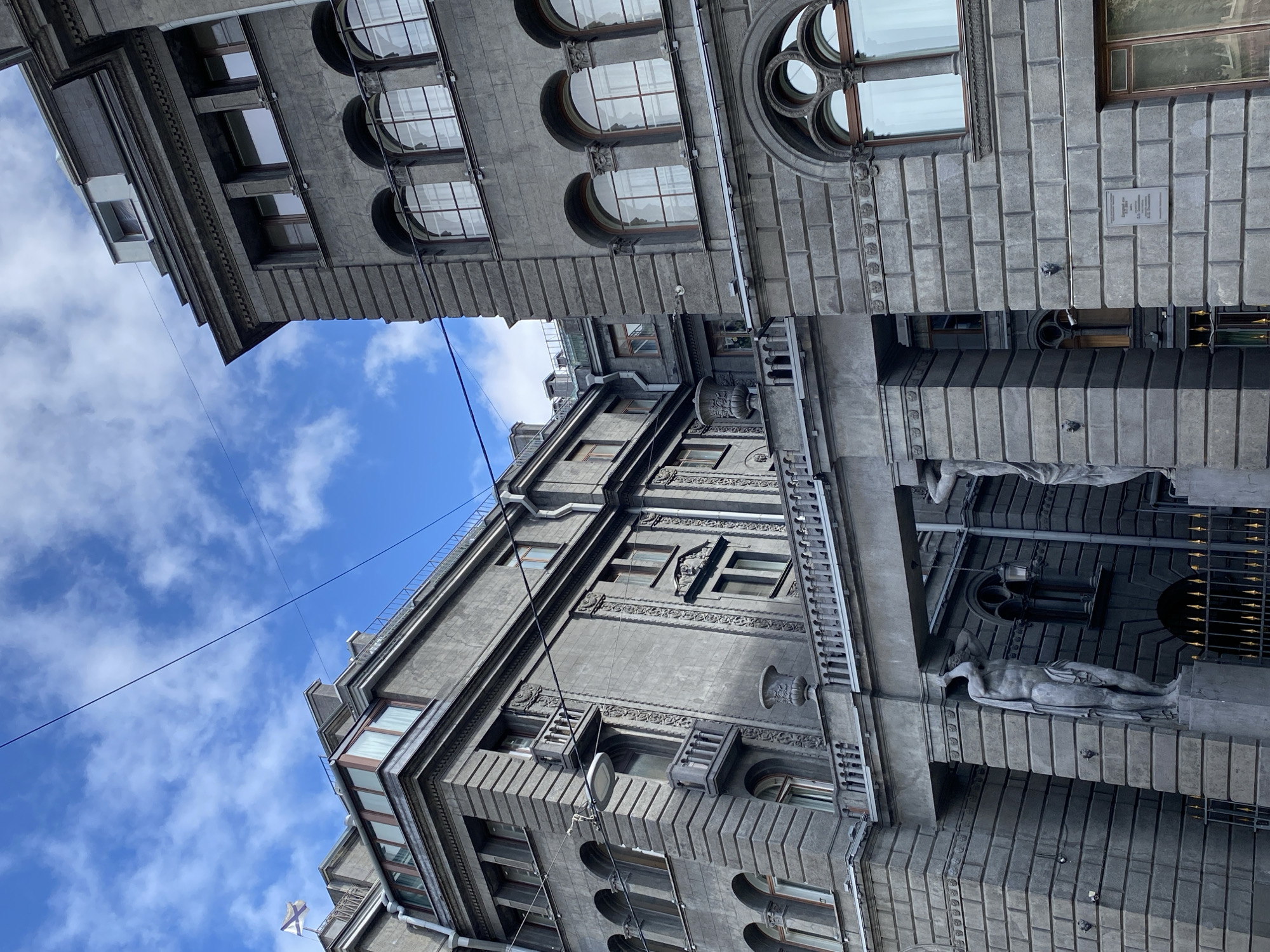 This image captures a view of the Singer House in Saint Petersburg, Russia, a prominent Art Nouveau building located on Nevsky Prospekt, overlooking the Kazan Cathedral. The photo focuses on the architectural details of the building's facade, characterized by its imposing grey stone construction, numerous windows, and ornate decorative elements.
The left side of the image showcases a section of the building with a balcony supported by classical statues, one of which is prominently visible. This architectural feature suggests an entrance or a significant decorative element. Further to the left, another part of the building rises, featuring a series of arched windows and a flag with a distinctive pattern flying from a rooftop.
The right side of the image presents a more compact view of the Singer House, highlighting its upper floors. Here, a series of large, arched windows with decorative frames dominate the facade. The stonework is finely detailed, with rows of rectangular blocks and intricate cornices. A plaque is visible on the stone facade.
The sky above is a bright blue with scattered white clouds, suggesting a clear day. Two dark wires, likely power or telephone lines, stretch across the frame, intersecting the buildings and the sky, adding a modern element to the historical architecture. The lighting suggests it is daytime, with shadows cast by the architectural elements. No people are visible in the frame. The overall impression is one of grandeur and historical significance, with a focus on the intricate design and imposing scale of the Singer House.