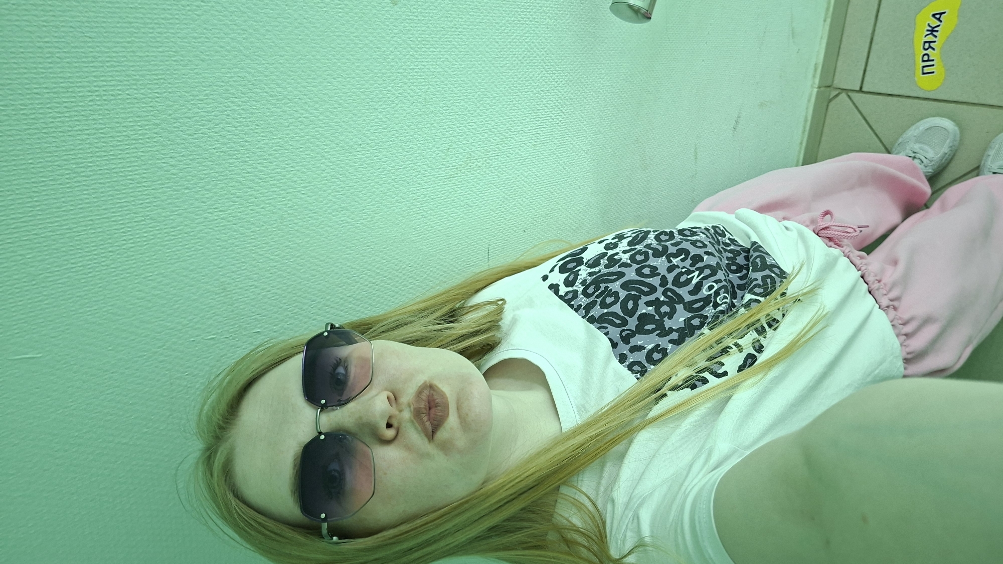 A woman with long blonde hair is lying down, wearing sunglasses, a white t-shirt with a leopard print patch, and pink sweatpants. She is indoors with a light green wall and tiled floor. A sign that reads "ПРЯЖА" in yellow and black Cyrillic letters is visible on the floor tiles. The location is likely Chaikovsky, Russia, based on the Cyrillic text. There are also a pair of tennis shoes. A showerhead is also visible in the top portion of the picture.