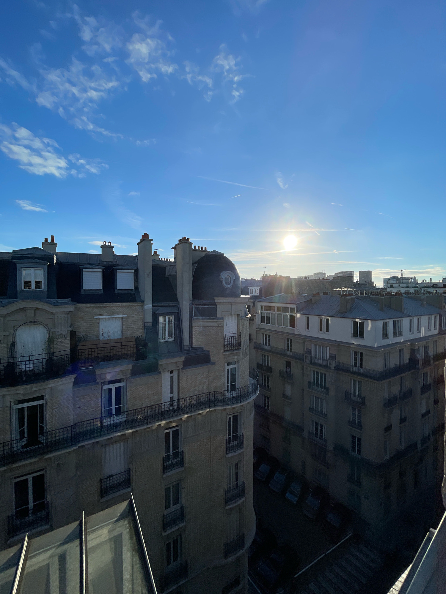 The image shows a view of Parisian rooftops under a clear blue sky with scattered clouds. The sun is shining brightly, creating lens flares. The architecture is characteristic of Paris, with mansard roofs, dormer windows, and ornate building facades. Several buildings are visible, some with chimneys and balconies. In the foreground, there is a modern glass structure, possibly a rooftop extension or balcony. The background shows a cityscape with more buildings and some construction cranes. Cars are visible parked on the street below. The lighting suggests it is either morning or late afternoon.