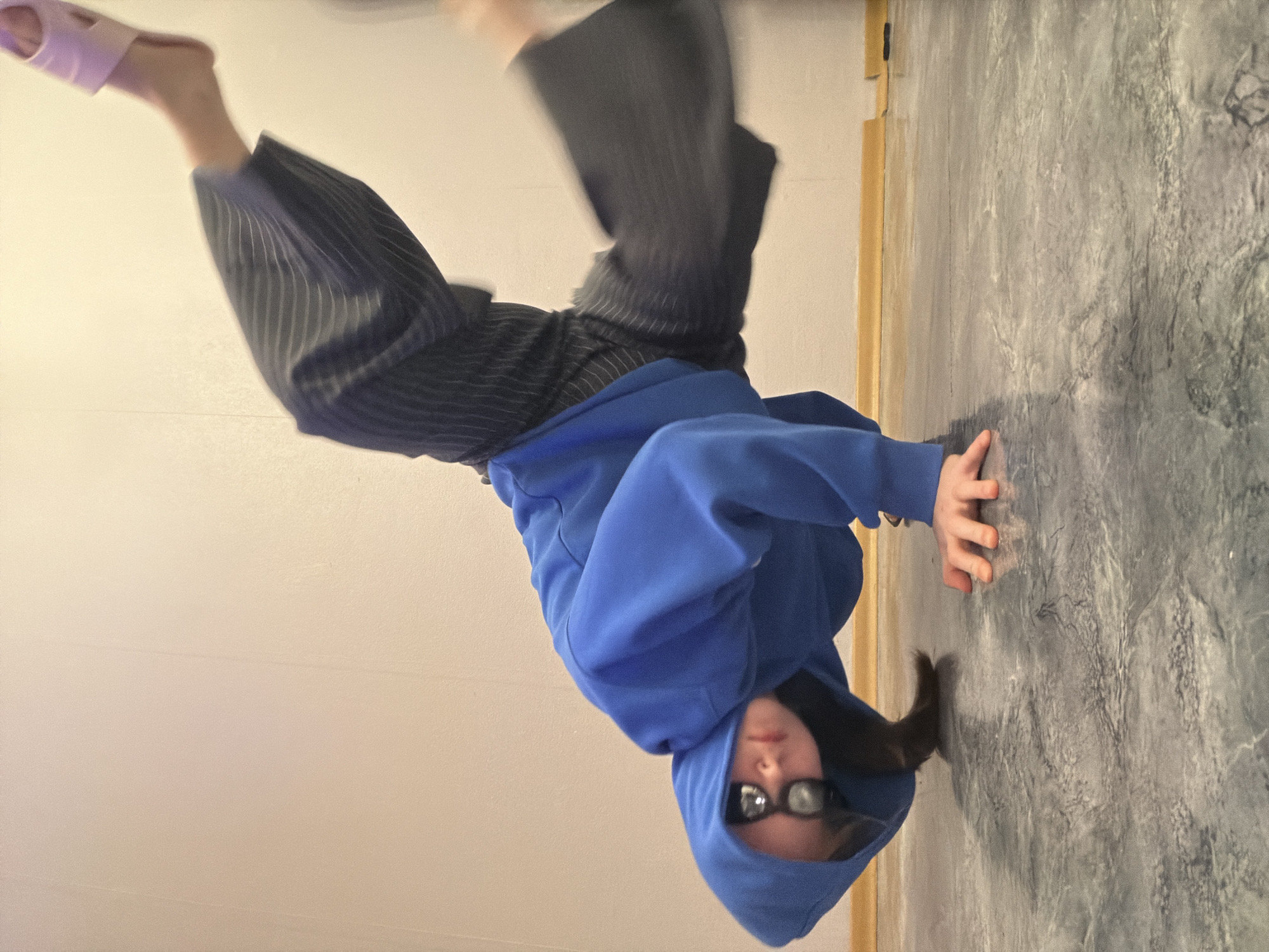 An indoor portrait-oriented image captures a person performing an inverted pose, likely a handstand or headstand, in Perm, Russia. The individual is positioned with their head near the bottom of the frame, facing upwards, and their legs extended towards the top. They are wearing a vibrant blue hooded sweatshirt with the hood drawn up, partially concealing their face, and distinctive dark-rimmed rectangular glasses. Their lower body is dressed in dark, pinstriped pants. One foot has a light pink slipper on it, while the other foot appears bare. Motion blur is visible on the person's legs and feet, indicating dynamic movement or the holding of a pose. The setting features a plain, light-colored wall on the left side, against which the person's feet are raised. To the right, a textured, dark gray surface, possibly a floor or another wall, serves as the support for the person's hands. A wooden strip runs vertically, separating these two background elements. The scene suggests a casual environment, possibly a home or studio. No text is visible in the image.
