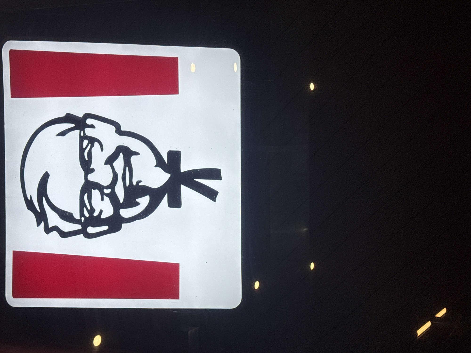 The image shows a lit-up sign for Kentucky Fried Chicken (KFC) at night. The sign features the iconic Colonel Sanders logo in black and white, set against a white background with vertical red stripes on either side. The sign is illuminated, and there are some blurred yellow lights visible in the dark background, suggesting ambient lighting or reflections. The bottom part of the image is dark, showing a textured surface that could be a wall or pavement with some subtle lighting effects. The overall impression is of a commercial establishment at night.