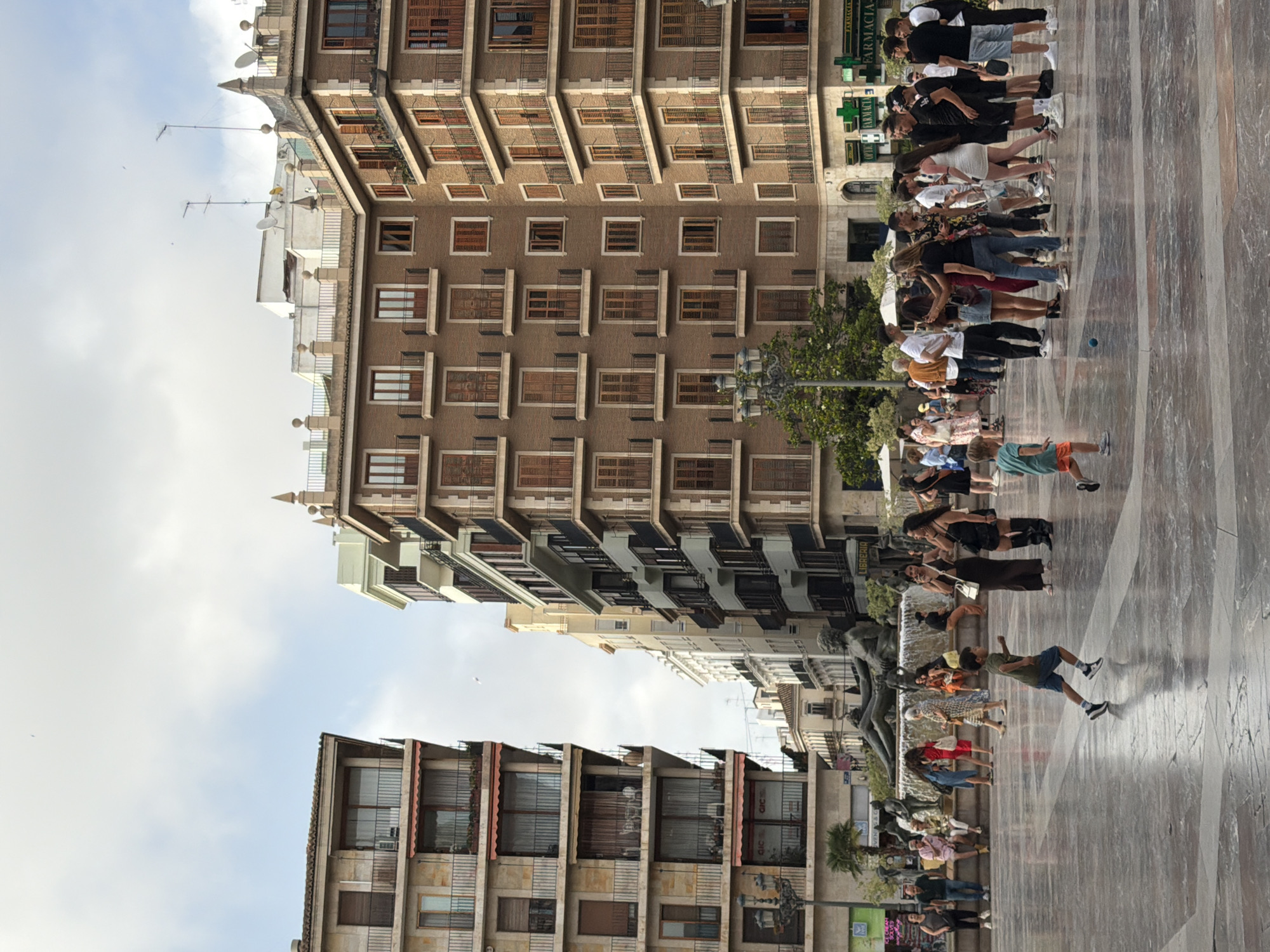 The image depicts a wide, paved public square in front of several multi-story buildings. The buildings are a mix of architectural styles, with some featuring ornate balconies and shutters, while others have a more modern facade. In the background, a large fountain with multiple sculptures is visible. A crowd of people are gathered in the square, some standing and talking, others sitting, and a few children are playing. The ground of the square is made of large, polished stone tiles with a marble-like pattern. The sky is partly cloudy. In the foreground, two young boys are running across the square. One boy, wearing a green t-shirt and blue shorts, is in motion with his arms outstretched. Another boy, wearing an orange t-shirt and blue shorts, is partially visible in the lower right corner of the frame, also running. A small blue ball is on the ground between the two boys. Several adults are present, some observing the children, others engaged in conversation. The overall atmosphere appears to be that of a lively, urban gathering space.