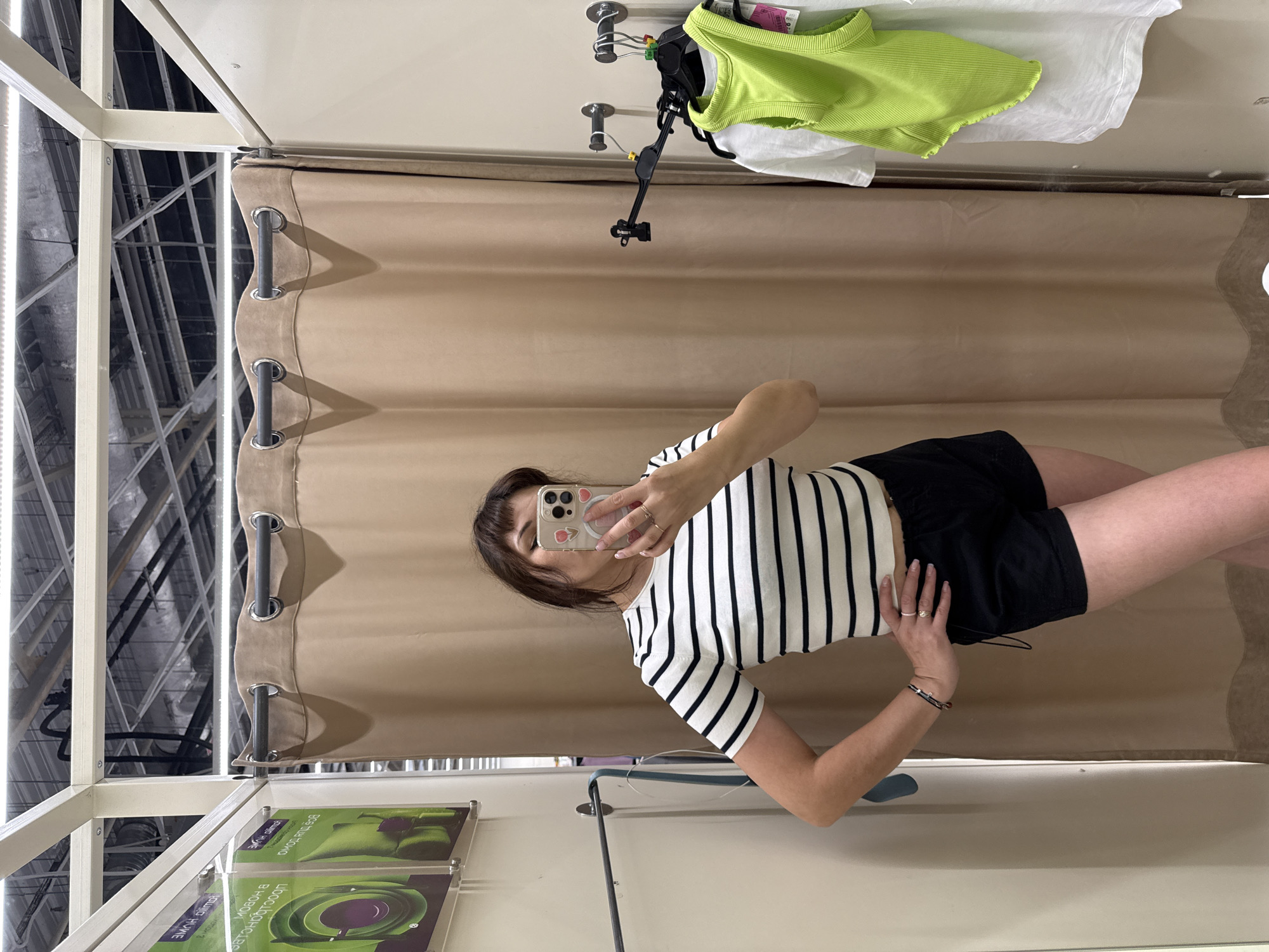 The image shows a young woman in a changing room, taking a mirror selfie. She is wearing a black and white striped t-shirt and black shorts. Her hair is dark brown with bangs, and she is wearing rings on her fingers and a bracelet on her wrist. She is holding a phone with a case featuring cartoon characters.
Behind her, a beige curtain hangs, typical of a fitting room. Above the curtain, a metal rod and hooks are visible. In the background, a rack of clothes can be seen, including a bright lime green top and a white t-shirt. The overall setting appears to be a retail environment, likely a clothing store. The lighting suggests it could be daytime, but there are no direct windows visible to confirm the weather or exact time of day. The image is captured from a slightly angled perspective, as if the user is rotating to get a full view of her outfit.