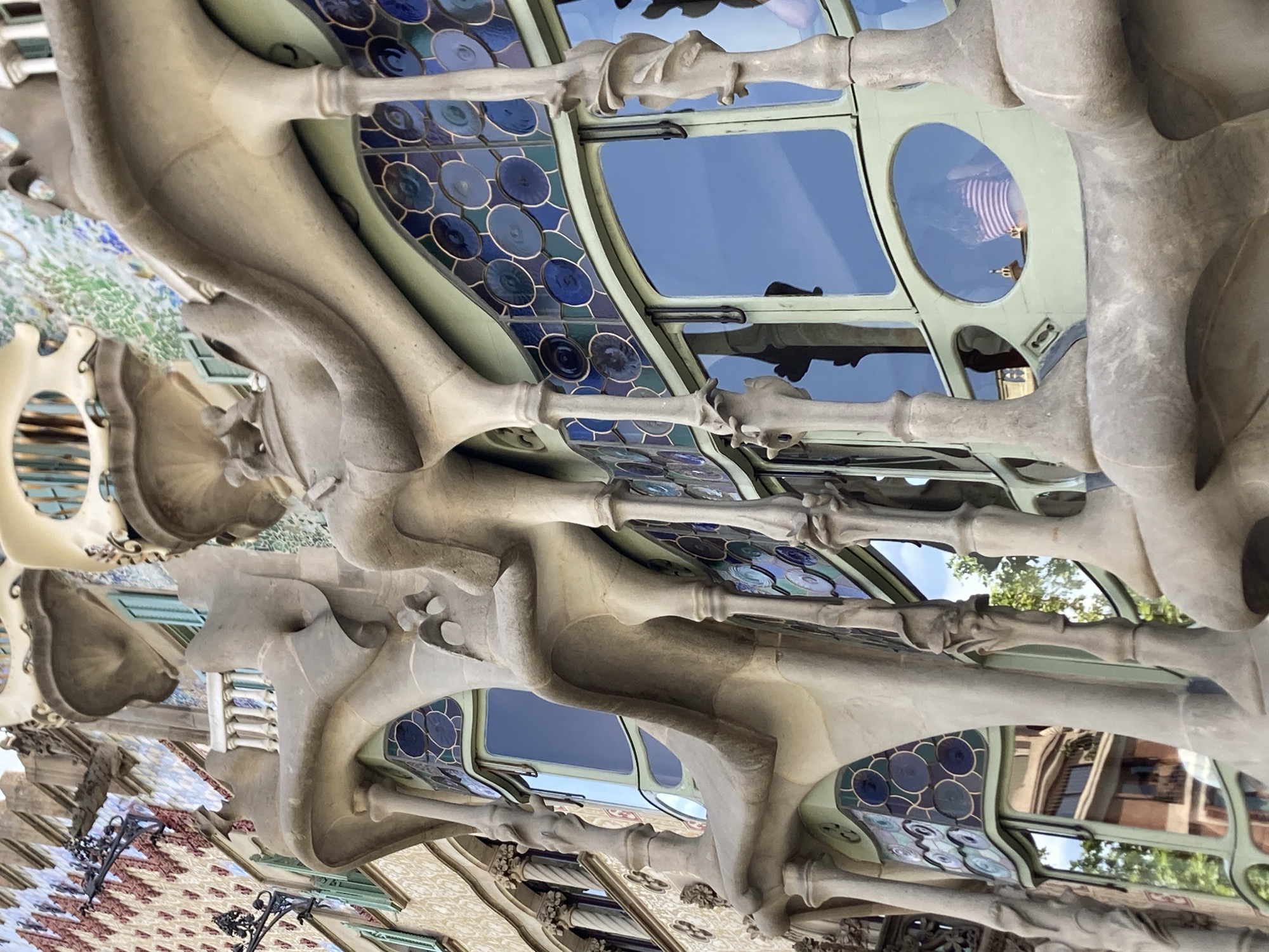 This is a close-up, low-angle photograph of the exterior of Casa Batlló, located on Passeig de Gràcia in Barcelona, Spain. The image showcases the building's distinctive Art Nouveau architecture, characterized by flowing, organic forms and vibrant colors.
The foreground is dominated by the building's facade, rendered in textured, light-colored stone that mimics bone or a skeletal structure. These elements form curved balconies and supports, creating an undulating visual rhythm. Behind the stone structures, large windows with a pale green frame are visible, some with decorative stained-glass panels featuring circular patterns in shades of blue and green.
Reflected in the windows, we can see glimpses of the sky, trees, and other buildings across the street, suggesting a daytime scene. In one of the oval windows, a person wearing a striped shirt is visible from the back, looking outwards.
The upper portion of the image reveals more of the building's facade, adorned with colorful mosaic tiles and ornate architectural details. Black wrought-iron fixtures, possibly sconces or decorative elements, extend from the wall. The overall impression is one of a whimsical and intricately designed building that stands out in its urban environment.