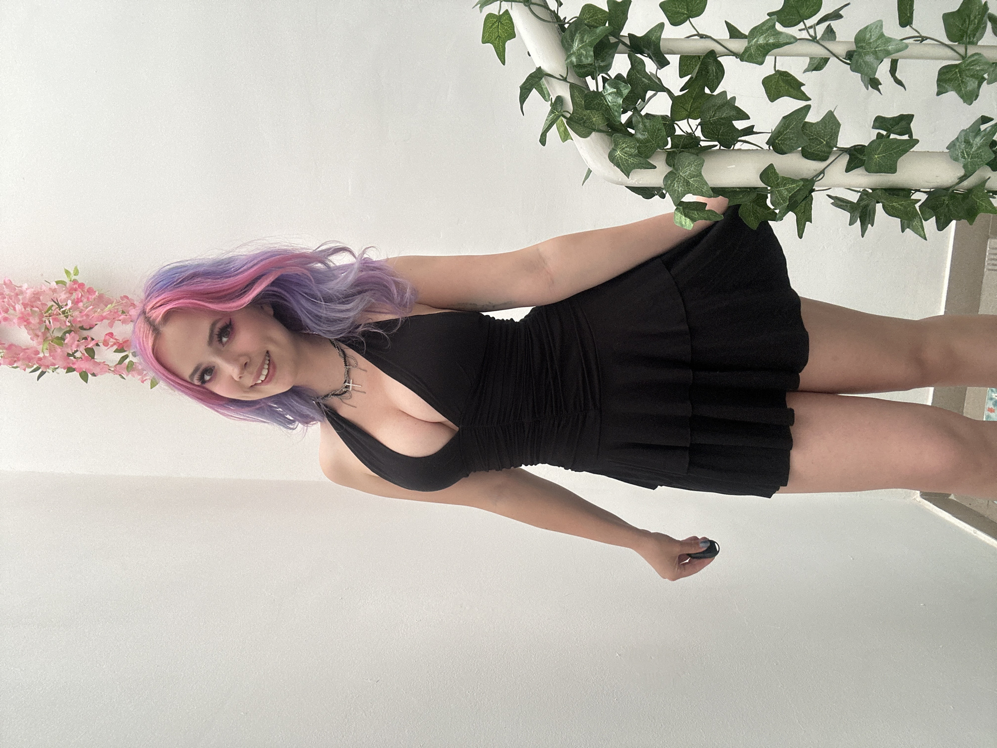 A young woman with long, wavy hair, vibrantly dyed in shades of pink and purple, is the central subject of the image. She is smiling warmly at the camera, conveying a cheerful and confident demeanor. She wears a black halter-neck mini dress with a ruched bodice that accentuates her figure and a short, flared, pleated skirt. A layered silver necklace adorns her neck. Her left hand is visible, holding a small, black object, possibly a remote or a compact device.
The setting appears to be indoors or in a well-lit, semi-enclosed space, characterized by plain white walls. To the left, a cluster of artificial pink flowers, resembling cherry blossoms, adds a decorative element. On the right, artificial green ivy is draped around a white railing or structure, suggesting a staircase or balcony area. The lighting is bright and even, indicating daytime conditions. There is no visible text within the image. While the context of the City of Zagreb, Croatia, is provided, no specific visual cues within the photo directly confirm this location.