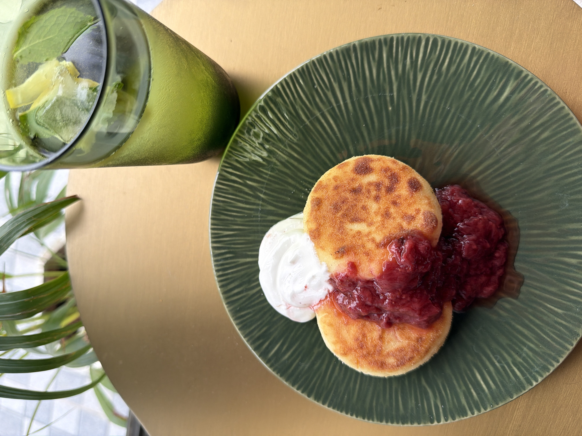 On a gold-toned surface, a green plate holds two small, circular pancakes topped with a dollop of white cream and a generous serving of red berry jam. To the right of the plate, a green glass contains a beverage with ice, mint leaves, and lemon slices. The background features a blurred green plant with long, slender leaves and a hint of an outdoor setting with a window. The scene is photographed from a high angle, focusing on the food and drink. The location is not identifiable from the image.