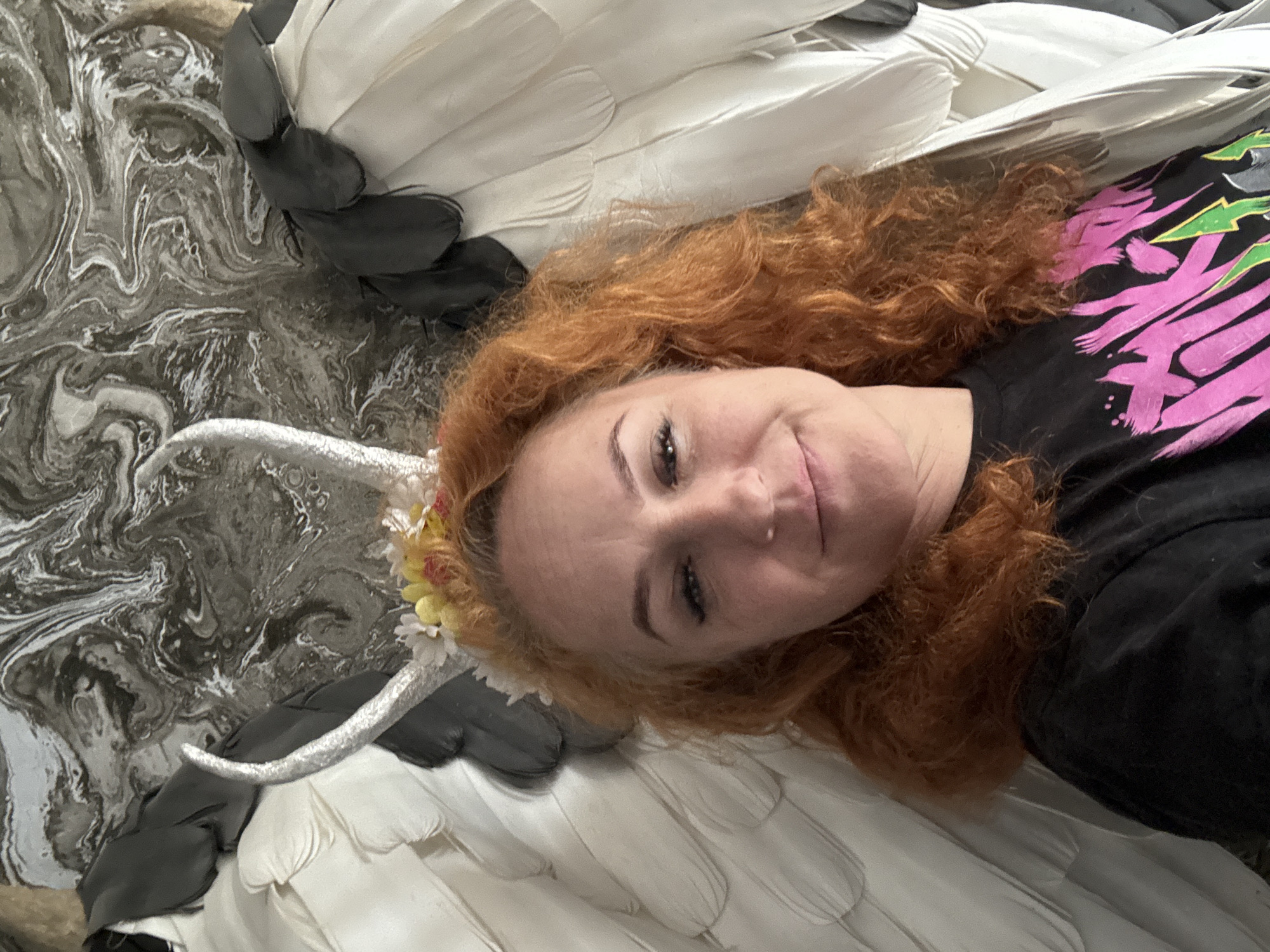 A person with red curly hair, wearing a floral crown with silver horns and a black t-shirt with a pink design, is lying down against a backdrop that includes white and black feathered wings and a marbled surface. The location is not specified.