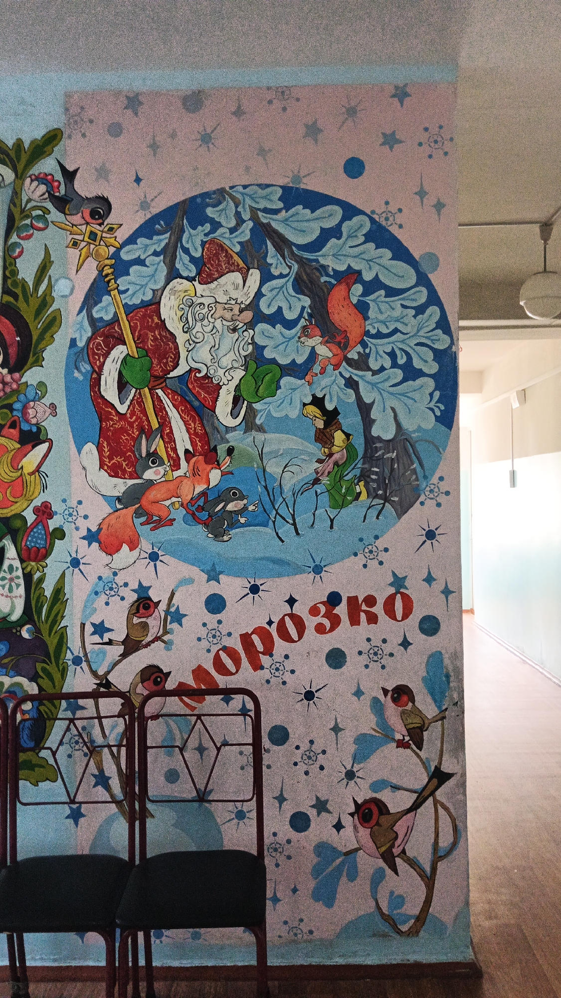A mural depicts a winter scene with Ded Moroz (Grandfather Frost) holding a staff and surrounded by woodland creatures, including a fox, rabbits, a squirrel, and a bird. The mural features a circular element with stylized branches and snow. Below the circular scene, the Cyrillic word "МОРОЗКО" (Morozko) is written in large red letters, interspersed with snowflakes and stars. The mural is on a light pink and blue background decorated with various winter motifs. Two dark chairs with metal frames are positioned in front of the mural. The scene appears to be indoors, with a hallway visible in the background, suggesting it is located in a public building such as a school or community center. The presence of the Cyrillic language strongly suggests the location is in Ukraine, and specifically, the reference to Ded Moroz and the style of the mural are consistent with Soviet-era children's art found in such institutions. The image was taken in Kryvyi Rih, Ukraine.