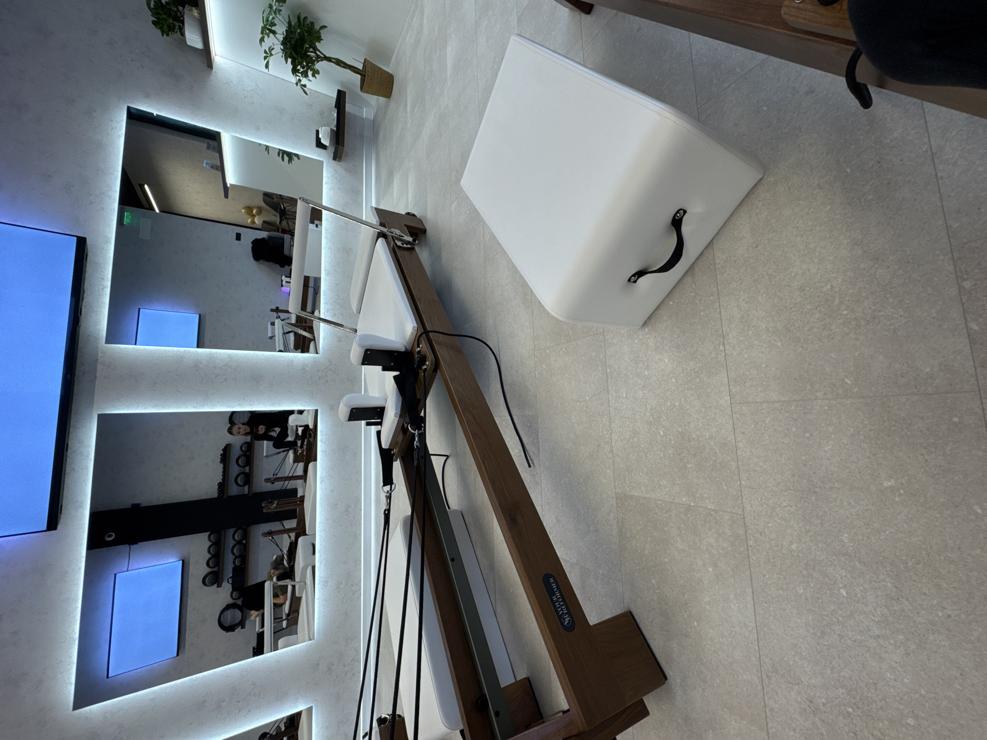 The image shows the interior of a modern fitness studio or wellness center, likely in Sofia, Bulgaria. In the foreground, there is a Pilates reformer machine made of wood with white upholstery. Next to it is a white cube-shaped ottoman or storage container with a black handle. In the background, a series of lit mirrors reflect the interior of the studio, showing exercise equipment, people engaged in activities, and additional screens. The overall color palette is neutral with white, gray, and wood tones. The floor appears to be made of light-colored tiles. There are plants on shelves near the mirrors, adding a touch of greenery.