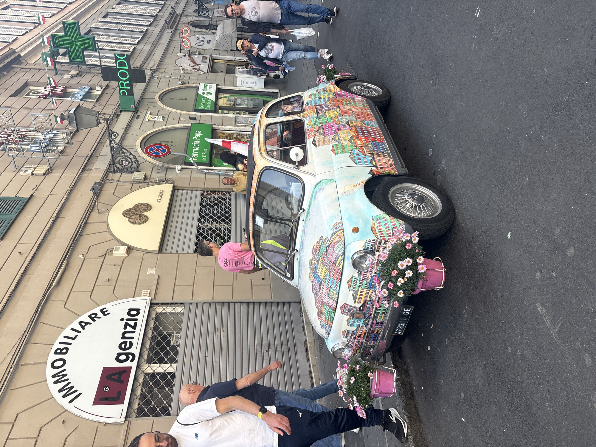 The image shows a street scene in Genoa, Italy. A brightly painted Fiat 500 car is parked on the street. The car's license plate reads "GE 479311 GE". The car is decorated with pink flower pots attached to the front and rear bumpers. On the left side of the car, a man in a pink shirt stands near the driver's side door. Two other men stand in front of the car, partially obscuring the view. Above and to the left, a sign for "IMMOBILIARE LA genzia" is visible. Further up, business signs for "Farmacia Papa Dr. E. Fazio", "Veterinariaalphega", "OTTICO", and "CAVO" are displayed on the building facades. Other people can be seen walking along the street.