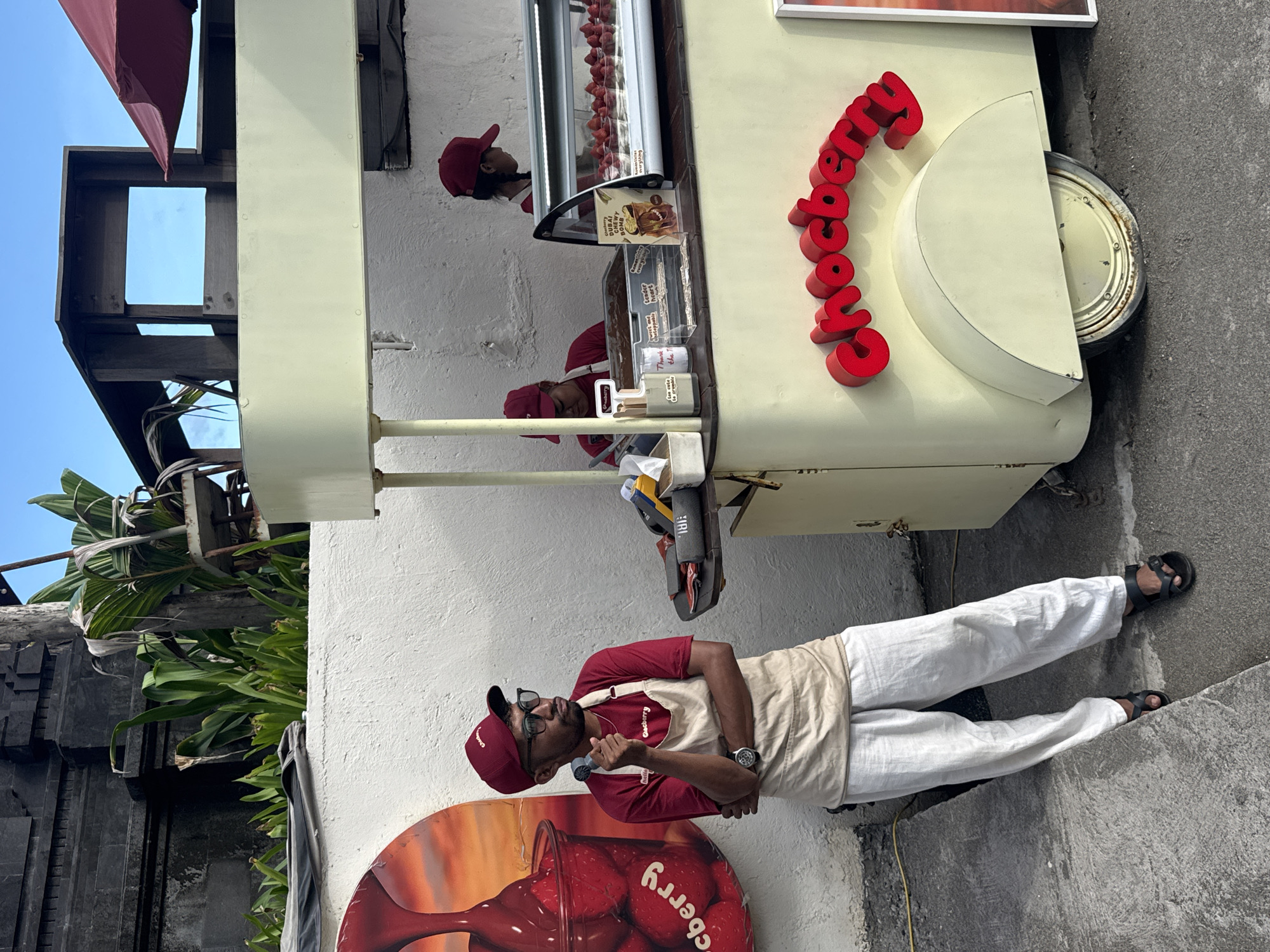 The image captures a street scene in North Kuta, Indonesia. A man wearing a maroon cap, glasses, and a maroon shirt under a beige apron is standing in front of a "Chocoberry" street vending cart. He is holding a microphone and appears to be speaking or announcing something. The cart is light yellow with the word "Chocoberry" in large red letters on the side. The cart displays items in a glass-fronted display case, including what appear to be strawberries and other confectionery items. The man is wearing white trousers and sandals. To the left of the cart, there are green plants and architectural stone details. The ground around the cart is a mixture of concrete and asphalt.