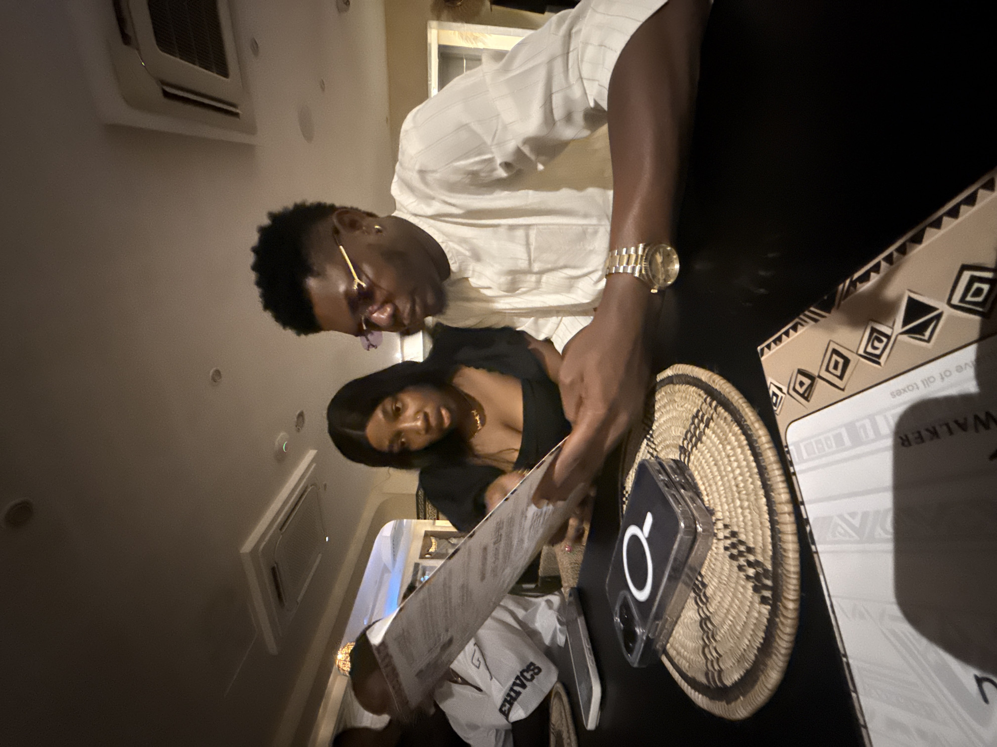 The image shows two individuals seated at a table in a restaurant, likely in Ikoyi, Nigeria. The man, wearing a white shirt and gold watch, is looking down at a menu. A woman in a black dress is seated next to him, also looking at the menu. On the table, there is a woven basket, a mobile phone in a clear case with a white logo, another mobile phone, and a Johnnie Walker branded object. The environment appears to be dimly lit, with air conditioning vents visible on the ceiling.