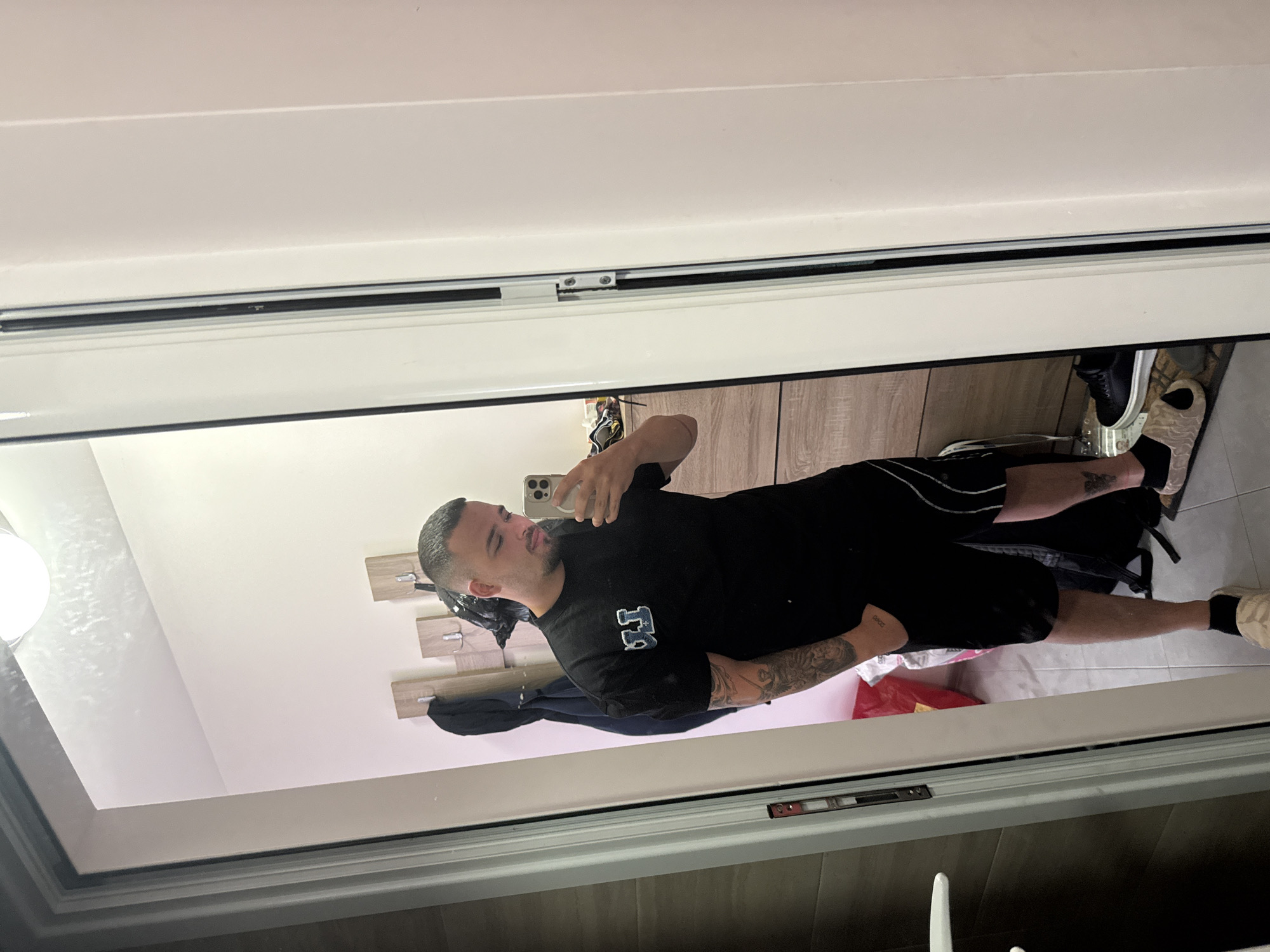 The image displays a mirror selfie of a man taken indoors, likely in Blagoevgrad, Bulgaria. The man has short, dark hair and a trimmed beard or stubble. He is wearing a black short-sleeved t-shirt featuring a light-colored "FJ" logo on the chest, black shorts, and light-colored sneakers with dark socks. Visible tattoos include a sleeve design on his right arm and a partially visible text tattoo on his left forearm that appears to read "3303."
He is holding a light-colored smartphone in his right hand, angled to capture his reflection in a large, framed mirror. The mirror's frame appears to be part of a door or wardrobe panel, with a white edge and a dark lining, showing some metal hardware. The background of the reflection reveals a bright indoor setting with white walls and a light-colored ceiling. On the left side, wooden panels with coat hooks are mounted on the wall, and a spherical light fixture is partially visible in the upper left. The floor appears tiled, and some clutter, including a red bag or clothing, is visible near his feet. The man's expression is neutral, as he is focused on his phone. The image is presented rotated horizontally.