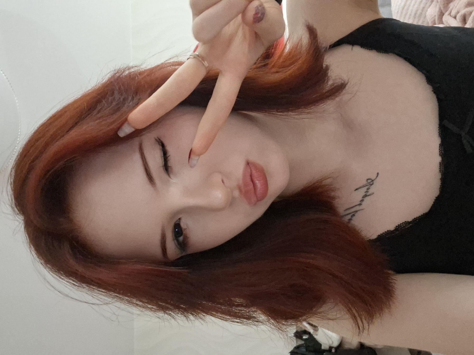 A young woman with red hair poses indoors, holding up her hand in a peace sign in front of her face. She is wearing a black top. A tattoo is visible on her upper chest. The location is implied to be the city of Chaikovsky, Russia.