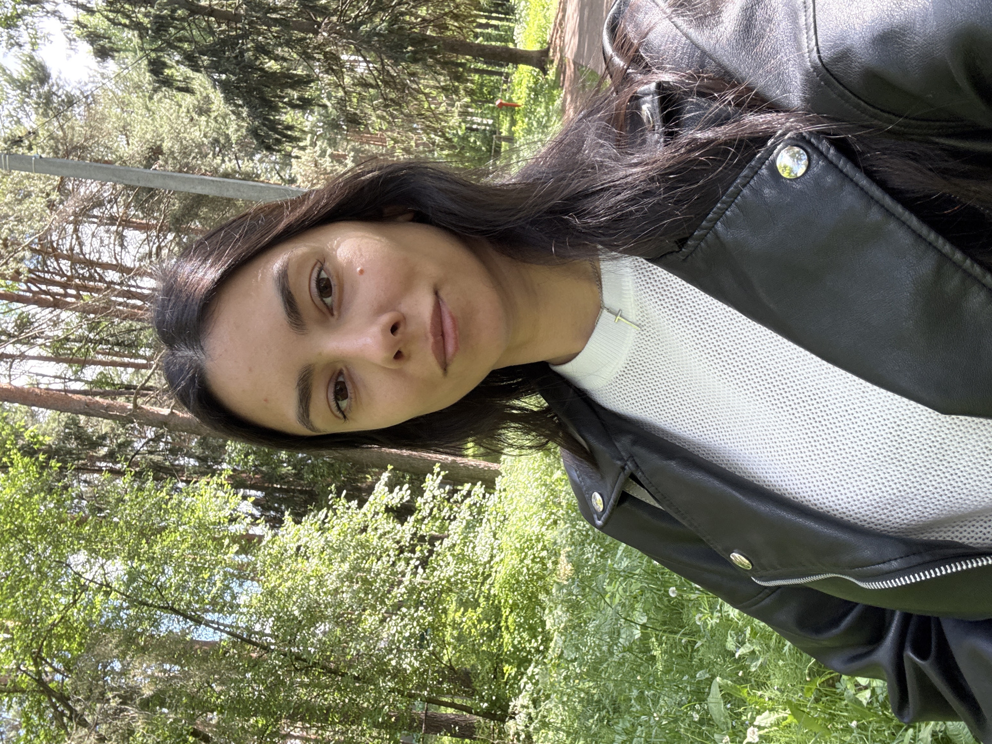 A woman with dark hair and medium complexion, wearing a white textured sweater and black leather jacket with silver buttons, stands in front of a wooded area with green undergrowth. The trees are various shades of green, typical of a forest or park. The lighting suggests it is daytime. This is the city of Chaikovsky, Russia.