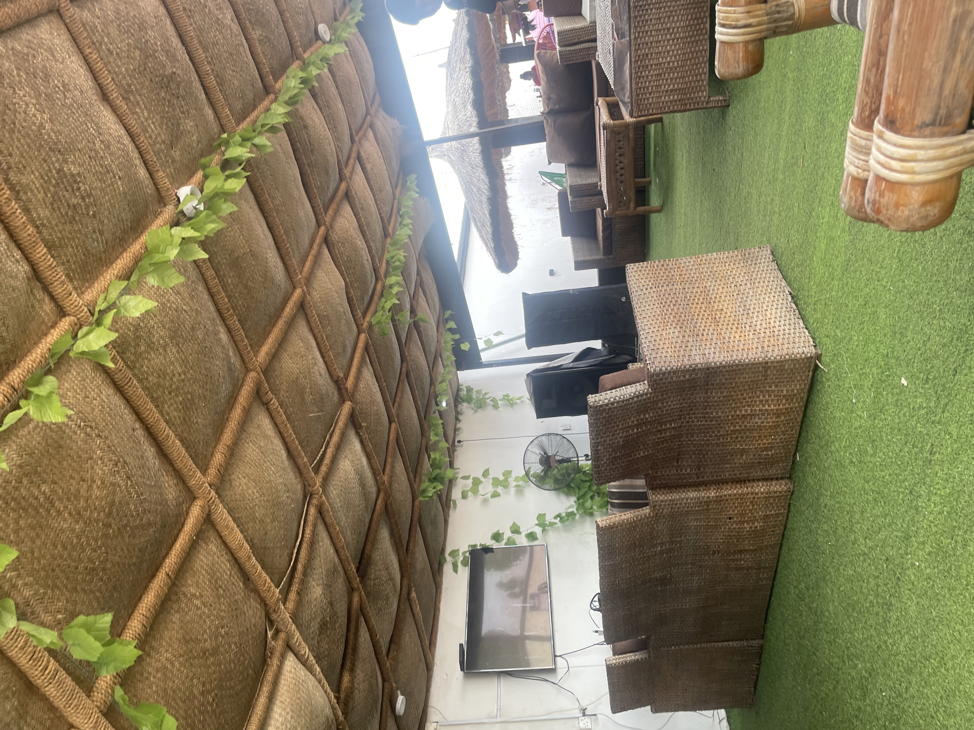 The image depicts an outdoor seating area with artificial grass flooring, likely a cafe or restaurant in Jauro Yinu, Nigeria. The scene is characterized by a thatched roof structure, with woven straw walls and a diagonal rope pattern. Artificial green vines with leaves are draped along the wall and across the ceiling.
Several pieces of woven furniture are visible, including armchairs and what appears to be a sectional sofa. These are made of rattan or a similar material. In the background, a thatched umbrella-style roof is prominent, suggesting an open-air dining or lounge space.
An electronic fan is mounted on the white wall, and a flat-screen television is also visible, mounted below the fan. Cables are seen connected to the TV. In the distance, through an opening in the structure, a few people can be partially seen, indicating that the establishment is likely open and operational. The lighting suggests it could be daytime, possibly late afternoon, with ample natural light. There are no discernible signs with text in the image. The overall impression is a relaxed, casual outdoor ambiance.