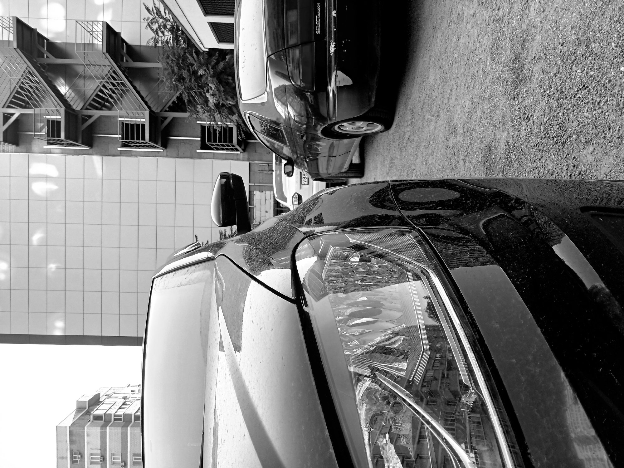 The image shows a close-up of the front of a black car, focusing on the headlight and hood. The car is parked on a gravel surface, possibly in a city environment. In the background, there are buildings, one with an external metal staircase, and two other cars are visible. The overall scene is monochromatic, suggesting a black and white photograph. The reflection in the headlight shows buildings with windows and trees, hinting at an urban setting. The image is likely taken in Krasnodar, Russia, given the context provided. There are no people or animals visible in the frame. The cars appear to be stationary, and there are no discernible actions or interactions taking place. The environment appears to be outdoors, with the sky visible at the top of the frame.