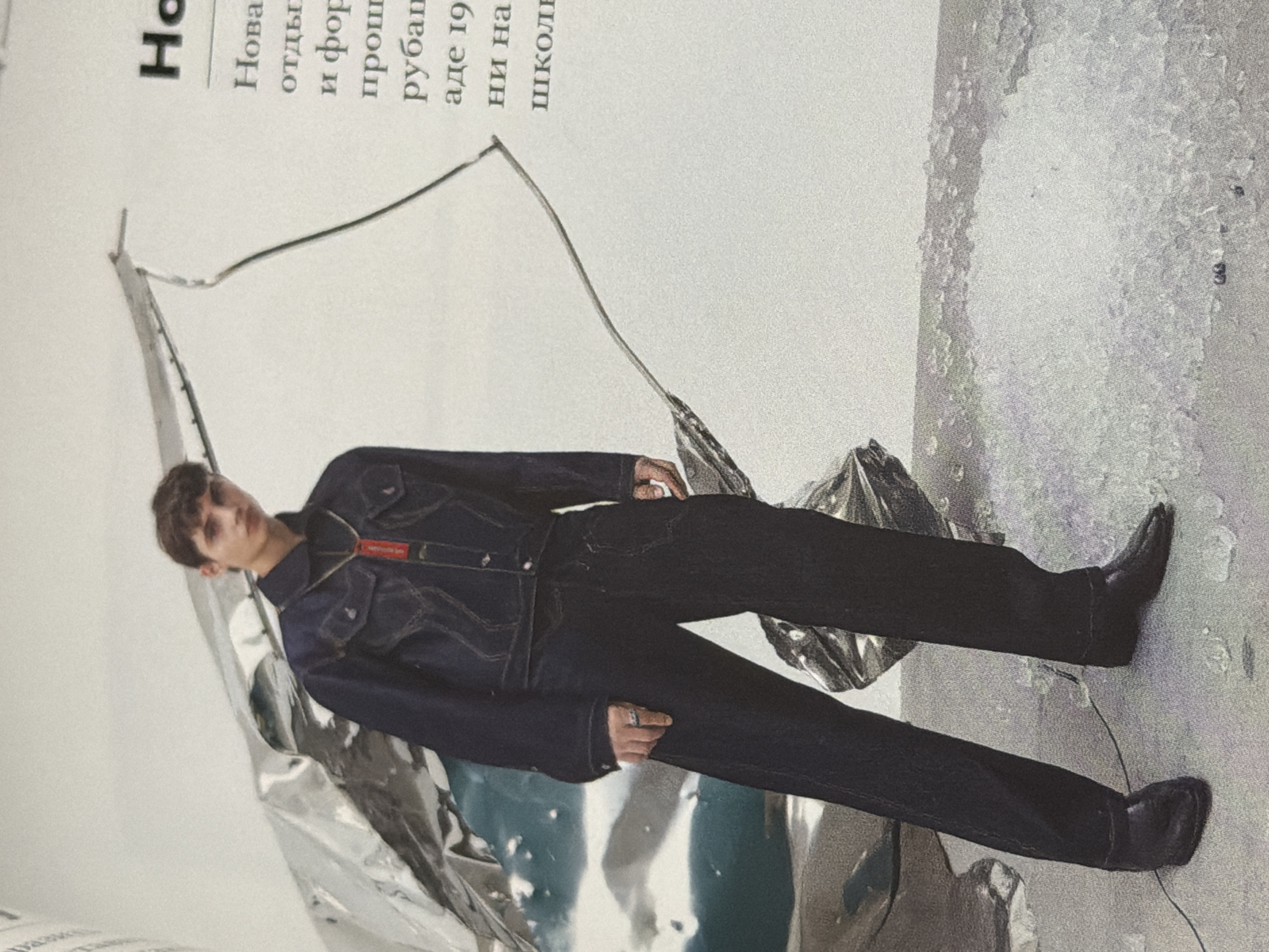 A fashion photograph depicts a male model posing on a magazine page, presumably for an advertisement or editorial. The model, seen from the chest down, is wearing a dark denim jacket and matching trousers, paired with black leather boots. He is positioned amid crumpled, reflective silver material, possibly intended to evoke a futuristic or abstract setting.
Text is visible in the upper left corner, written in Russian. The upper right corner features a textured white background.
Given the Russian text, it can be presumed that this photograph was taken in Russia. However, the photograph alone does not give sufficient information to make assumptions regarding its location.