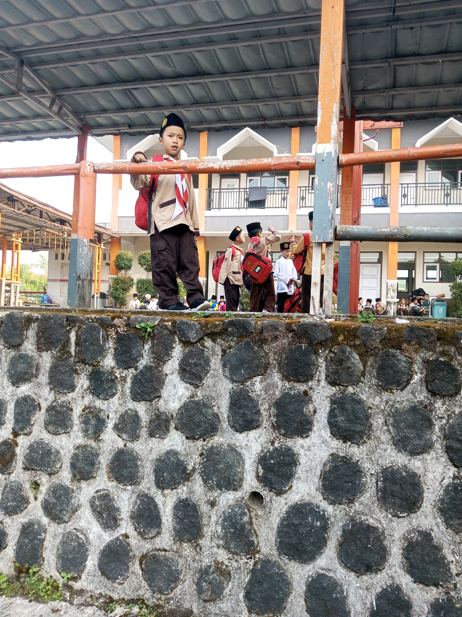 In Krasak, Indonesia, a young boy dressed in a school uniform stands on a stone wall. He wears a light brown uniform shirt, dark brown pants, a black cap, and a red backpack. Behind him, other children in similar uniforms are visible, some with backpacks. They are gathered in front of a building with multiple floors, orange and gray accents, and balconies. A covered walkway with a metal roof is also present. The stone wall is constructed with irregular dark gray stones and appears to be part of a drainage or retaining wall. Green vegetation is growing on and around the wall. The scene suggests a school or a gathering of students outdoors.