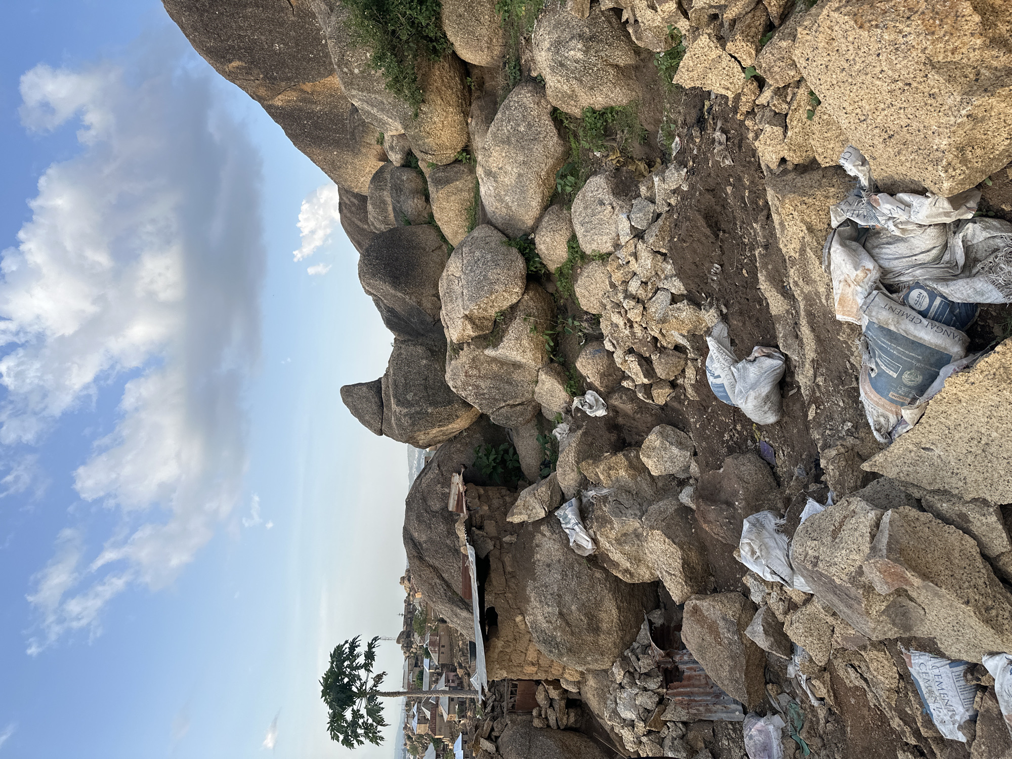 This image captures a rugged, rocky landscape under a bright, partly cloudy sky, characteristic of a natural environment in Jos, Nigeria. The scene is dominated by numerous large, weathered boulders of varying sizes, ranging in color from light brown to grey, which fill the foreground and midground. Sparse green vegetation, mainly grasses and small plants, grows between the rocks and in patches of exposed soil.
Scattered among the rocks are several white and off-white sacks. Many of these appear to be torn and discarded, with visible text on them, including "CEMENT," "MANGAL CEMENT," and "N.G.S.N," along with "52.5N" on one bag, indicating they are cement bags. This suggests human activity, possibly related to construction or refuse.
In the mid-distance, nestled among the rock formations, a settlement or part of a town is visible, with several buildings and structures. Some appear to be constructed with corrugated metal sheets, reflecting a more informal or utilitarian building style. A prominent, tall papaya tree with a full crown of leaves stands on the left side of the frame, providing a distinct botanical element against the rocky backdrop.
The sky is a clear blue with scattered white, cumulus clouds and a larger grey cloud to the upper left. The bright lighting suggests it is daytime, likely late morning or early afternoon. No people or active events are directly visible, presenting a still scene that highlights the interplay between natural geological features and human habitation in this distinctive environment.